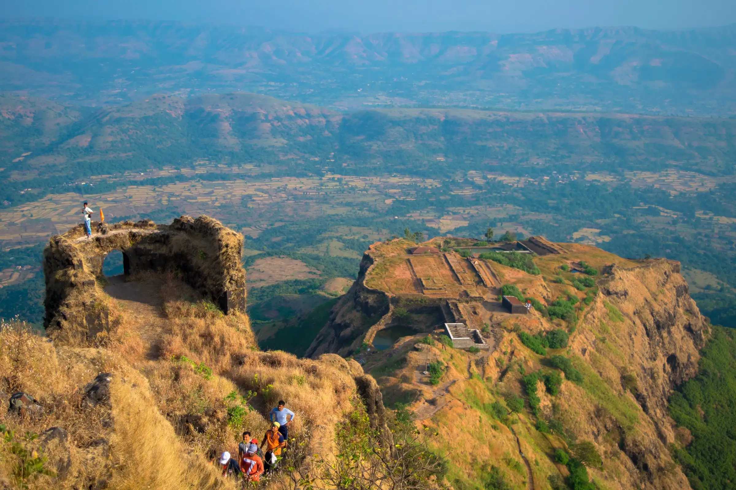 Trekkers climbing a grassy ridge on Rajgad Fort beside a ruined arched bastion, with a fortified machi and the farmed valley far below