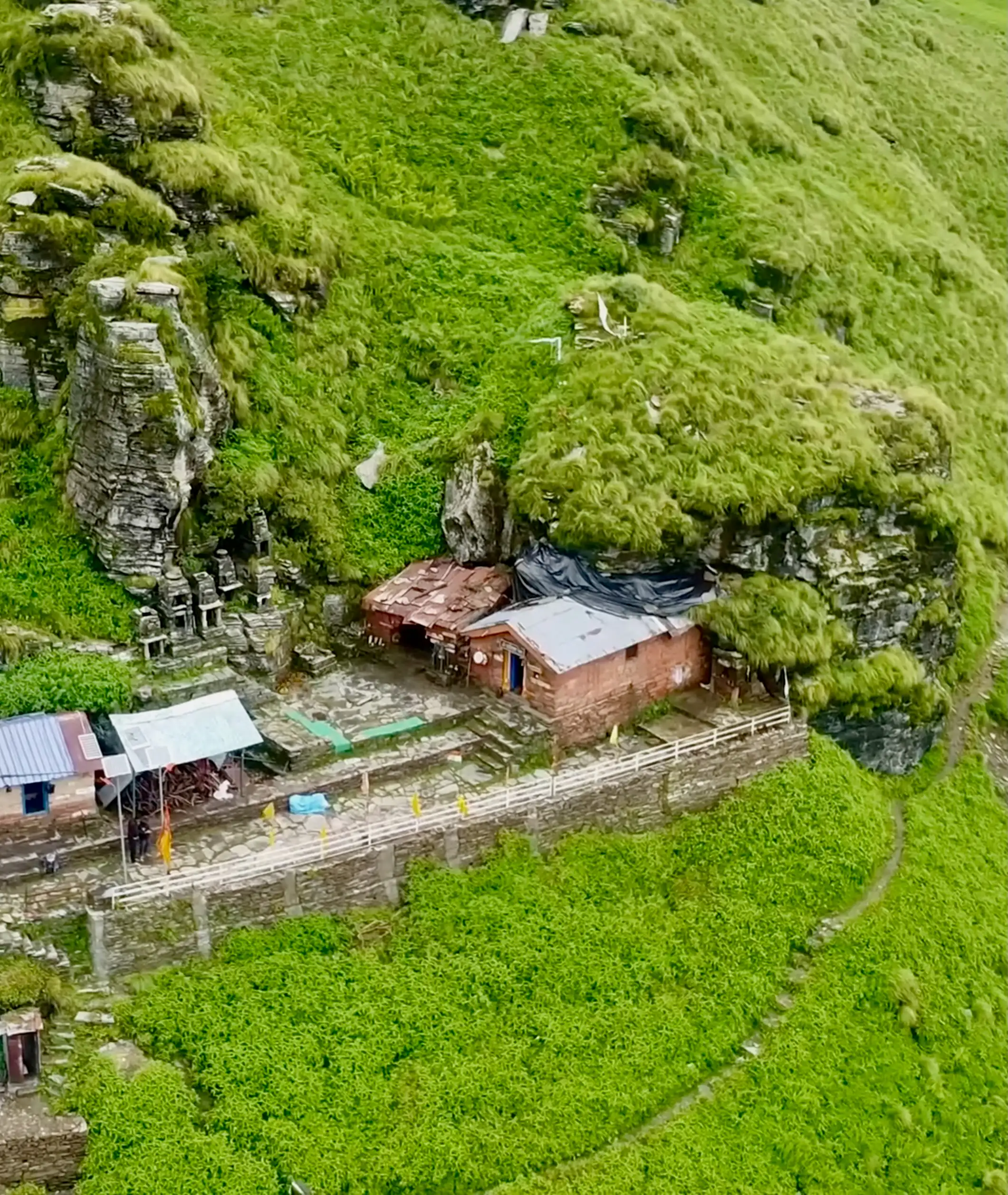 Aerial view of the Rudranath cave temple and its small stone shrines set into a green cliff, with the trail curving below