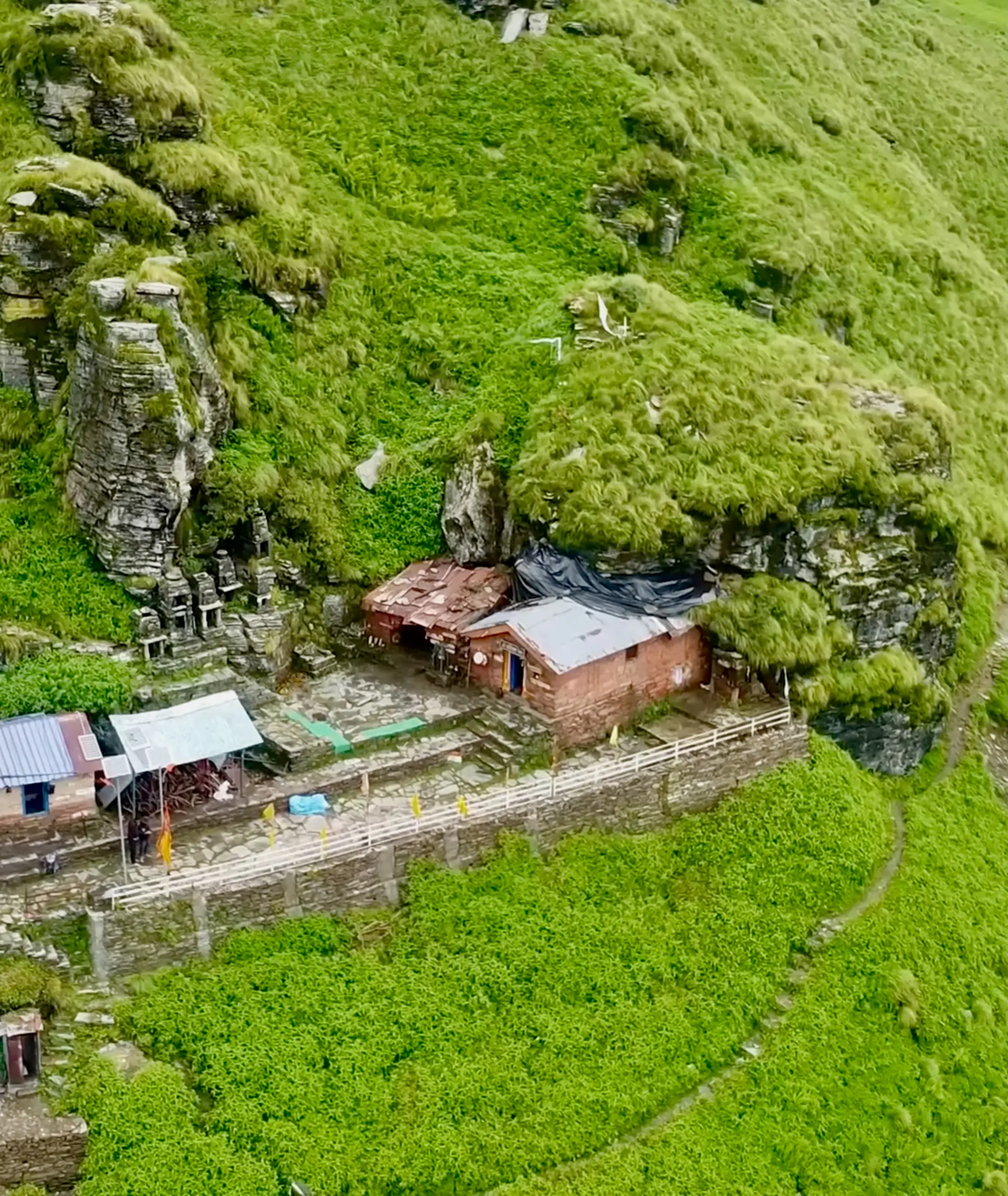 Aerial view of the Rudranath cave temple and its small stone shrines set into a green cliff, with the trail curving below