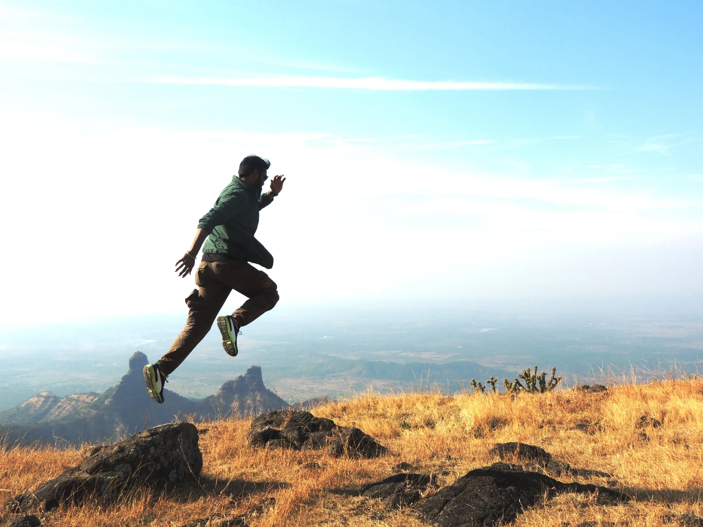 A trekker leaping across dry golden grass on the Ahupe plateau, with the twin rock pinnacles of Gorakhgad and Machindragad and the hazy Konkan plain far below