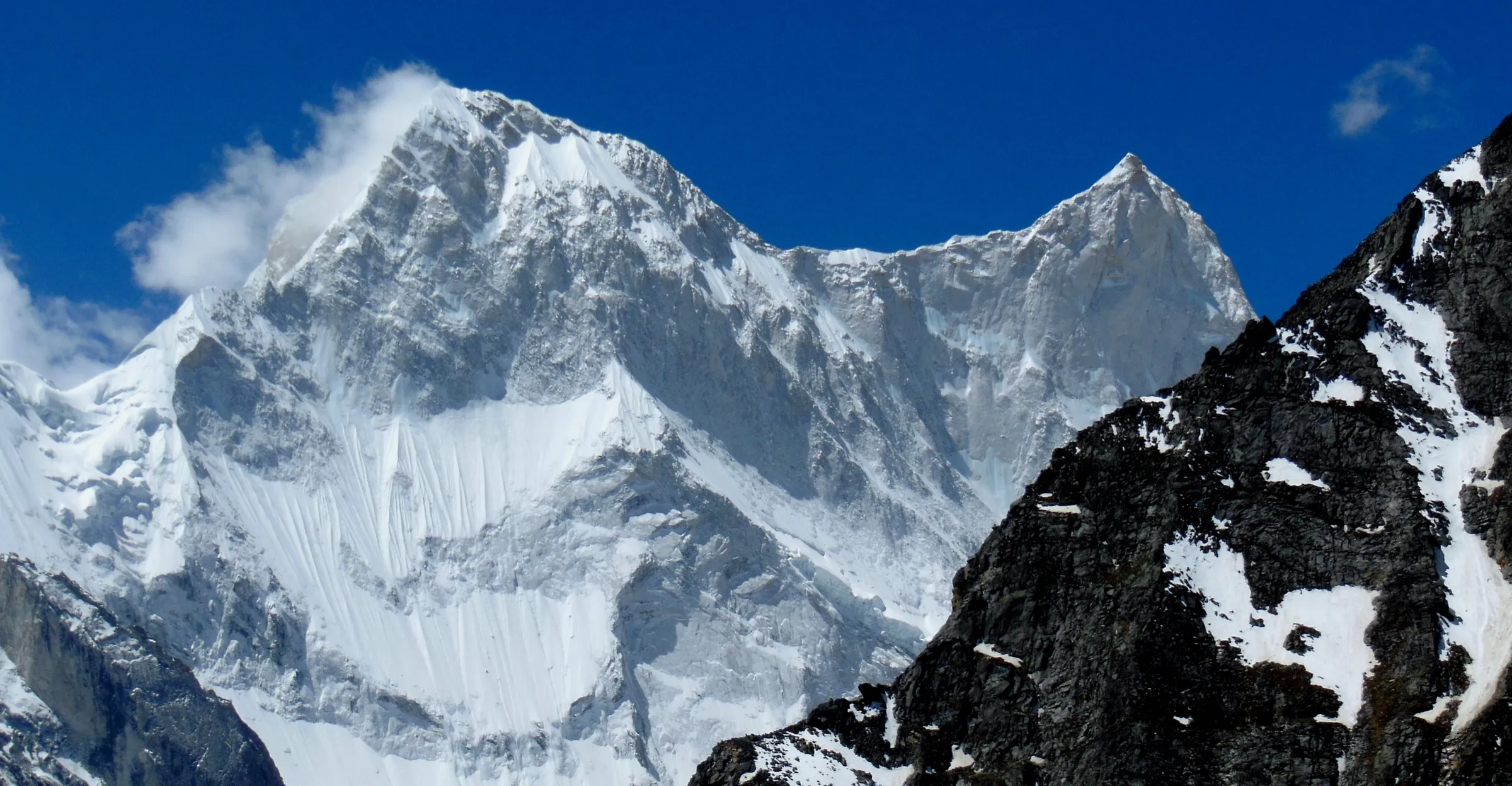 Snow-plastered Kalanka and the pointed summit of Changabang under a deep blue sky, seen from the Bagini Glacier with a dark rock ridge in the foreground