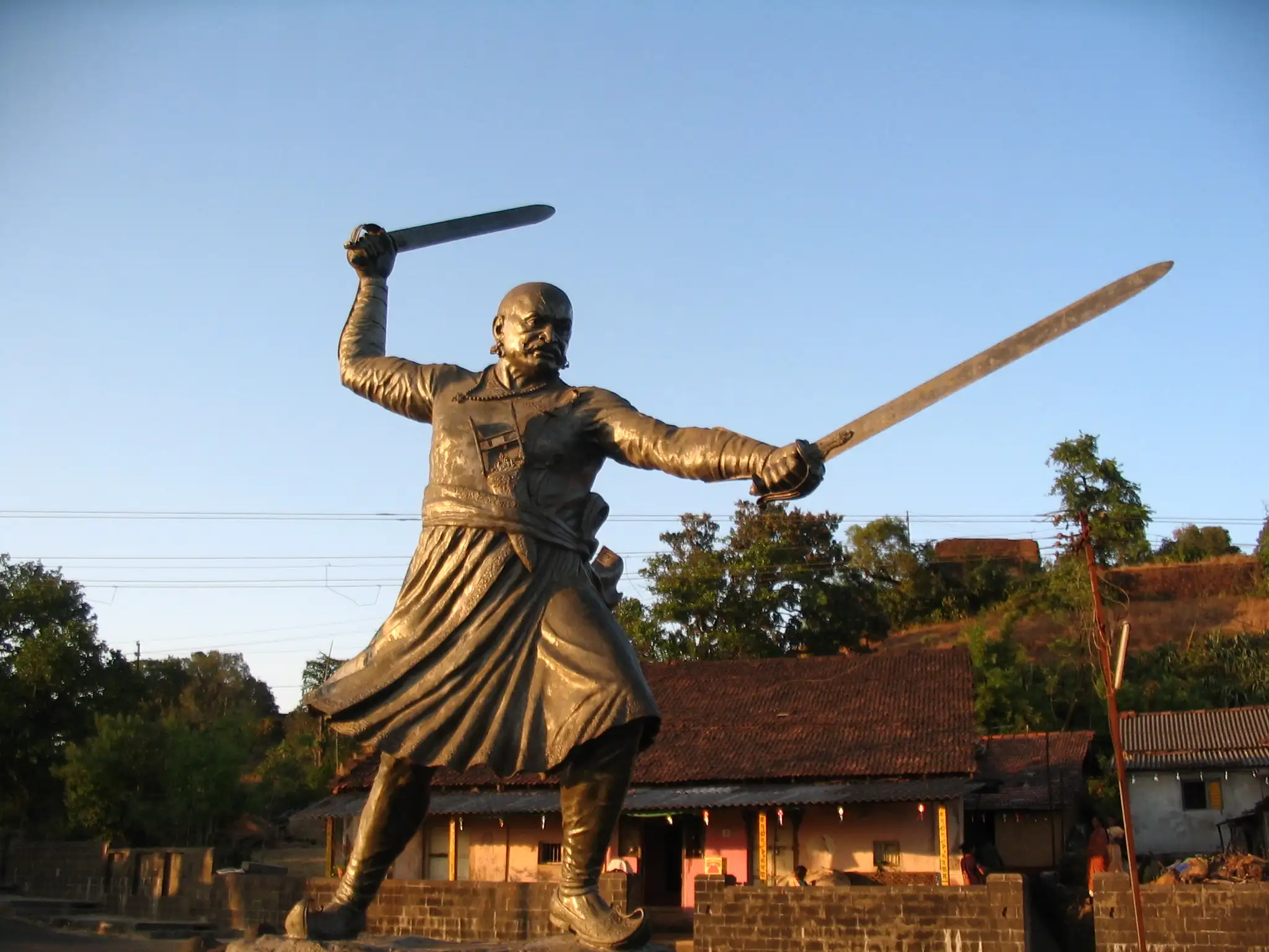 Bronze statue of Baji Prabhu Deshpande holding two swords, with tiled village houses behind it on Panhala Fort
