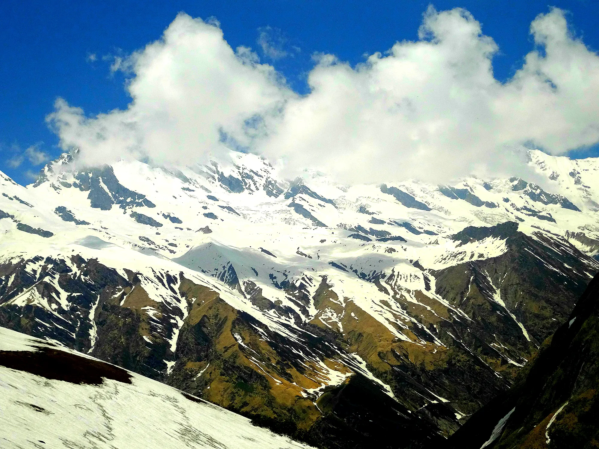 Snow-covered Himalayan peaks and steep green slopes under white clouds, seen from Baluni Top in the Sunderdhunga valley