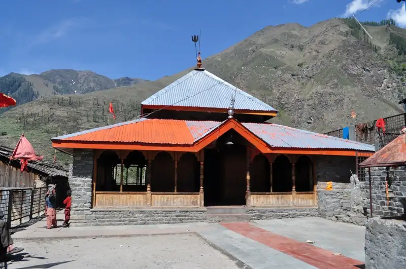 The Mahakali Banni Mata temple near Tundah, with an orange roof and red flags below green and brown mountain slopes; trekkers seek permission for the crossing here