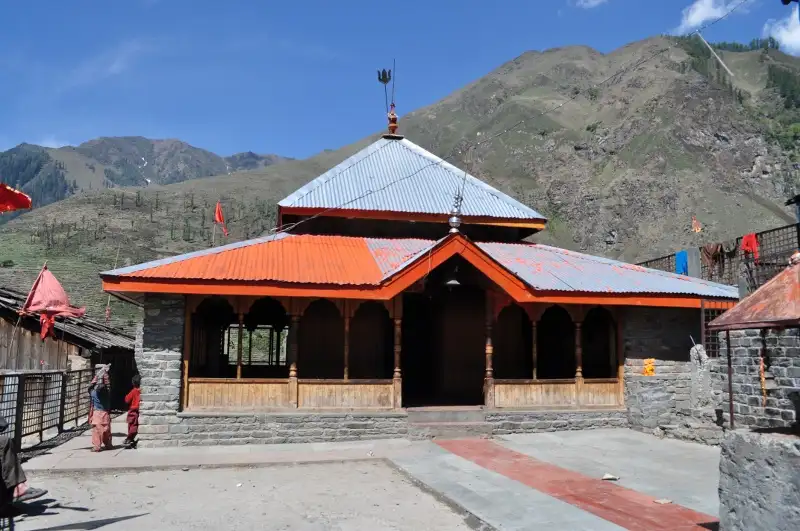 The Mahakali Banni Mata temple near Tundah, with an orange roof and red flags below green and brown mountain slopes; trekkers seek permission for the crossing here