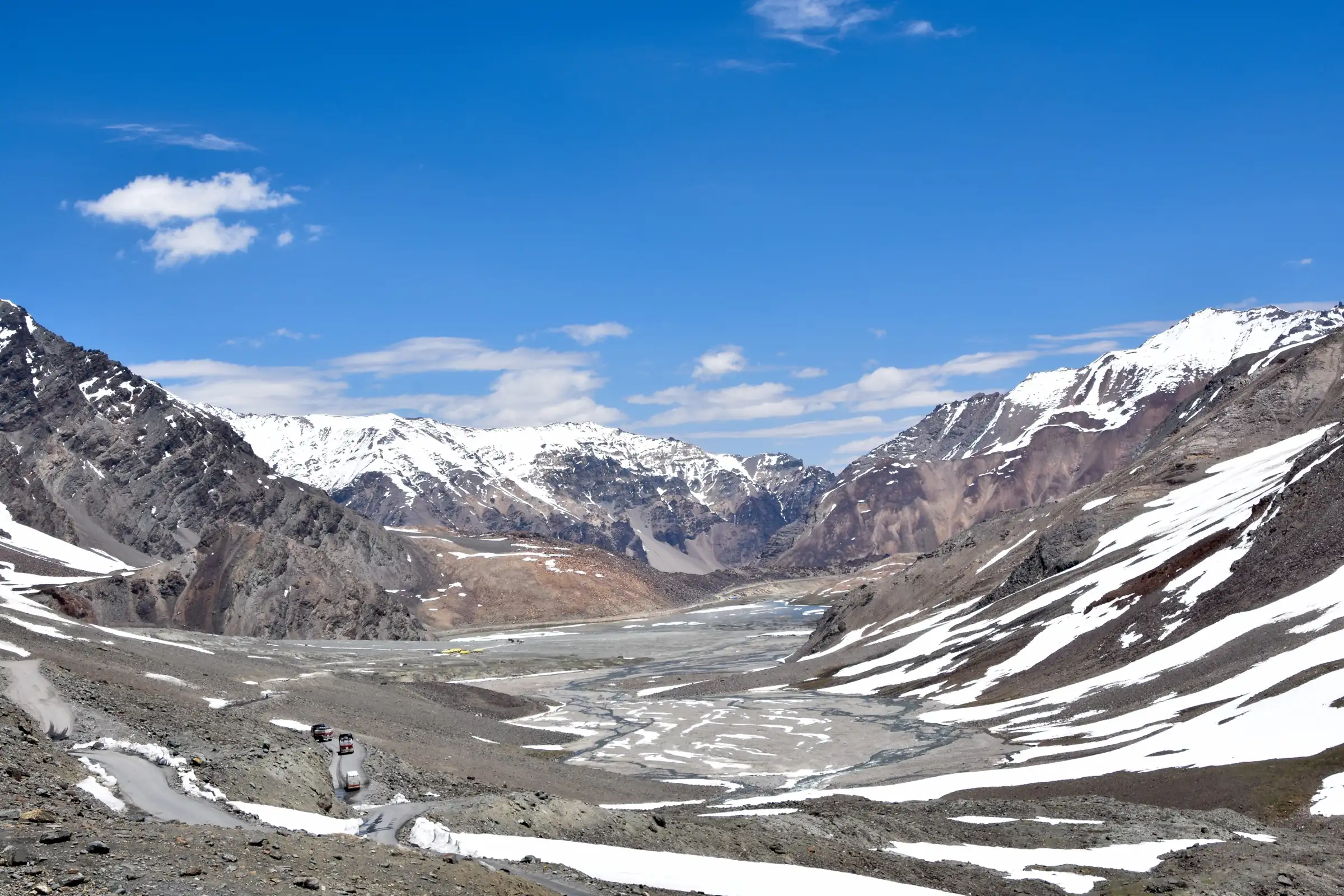 The Manali-Leh highway descending from Baralacha La into a wide snow-streaked valley, with the yellow tents of Bharatpur camp, the trailhead for Yunam Peak, on the valley floor