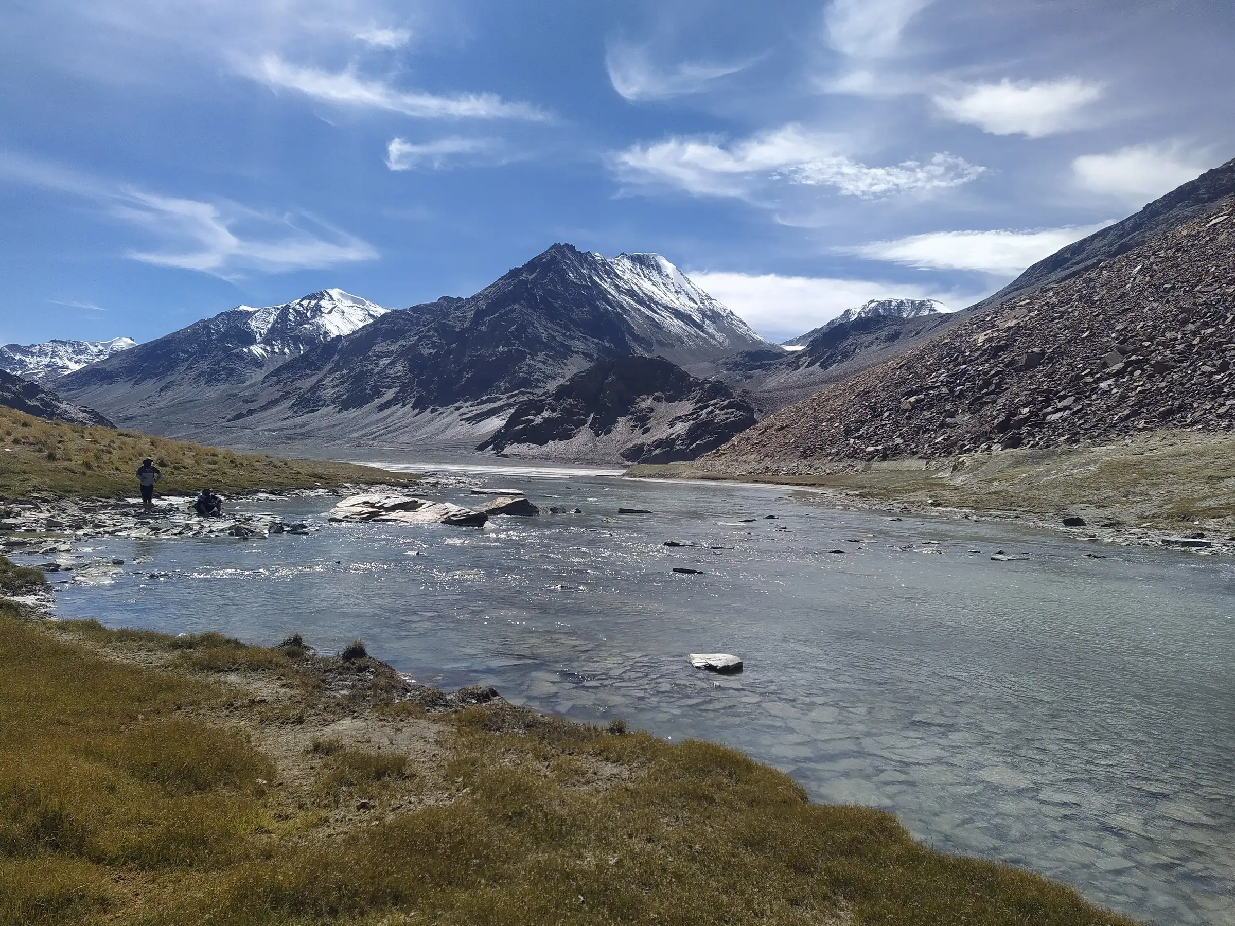 A shallow glacial river and grassy banks below snow-dusted peaks in the Baralacha La area, near the end of the trek