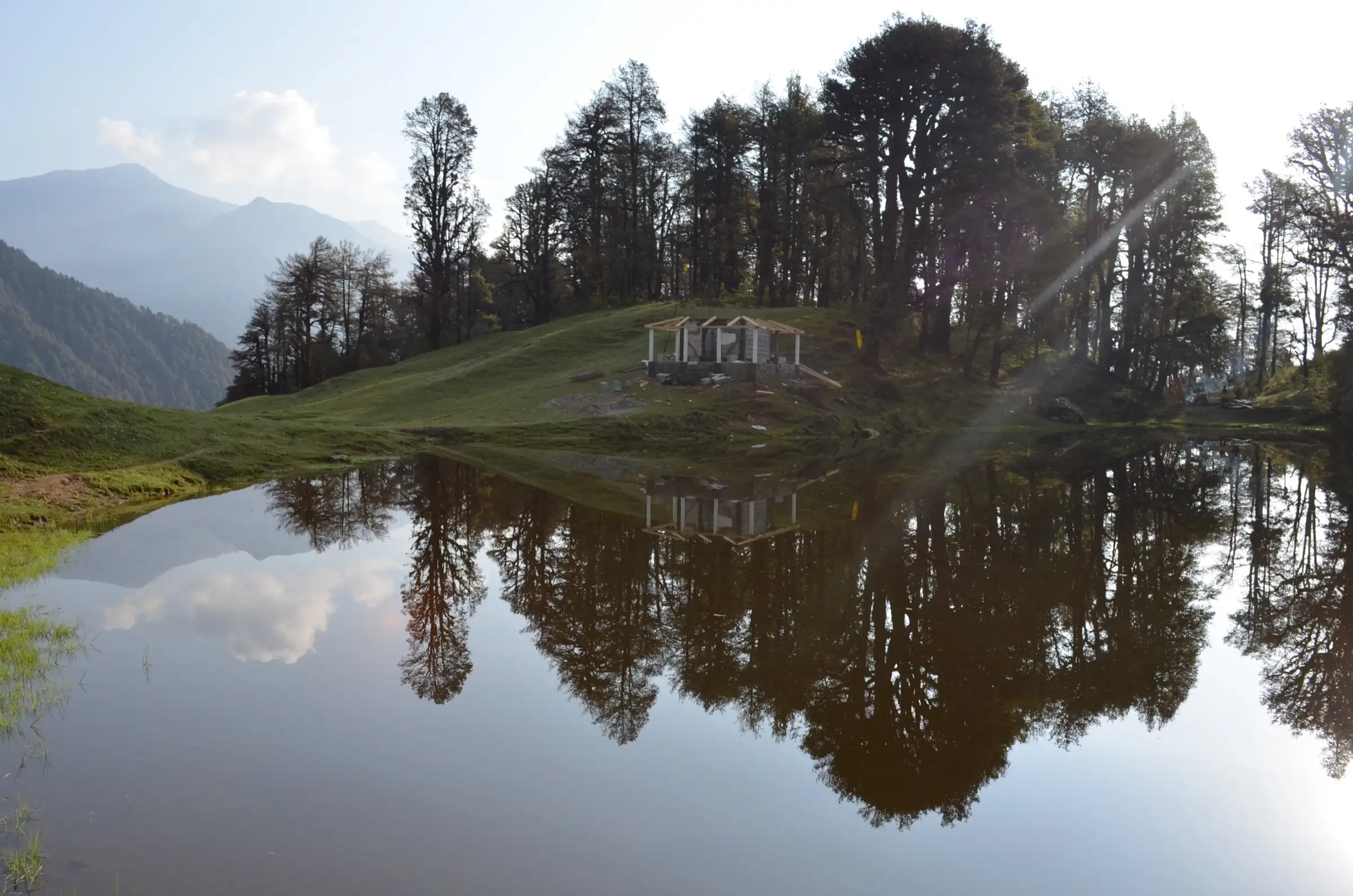 Barnala Tal lake reflecting tall trees and a small shelter, with mountains in the haze behind
