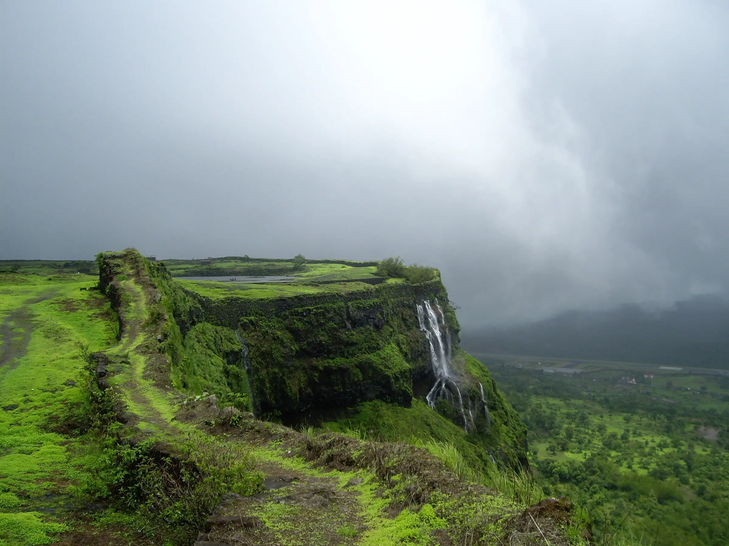 Green moss-covered rampart and bastion on the Korigad plateau, with a waterfall pouring off the cliff into the valley under monsoon cloud