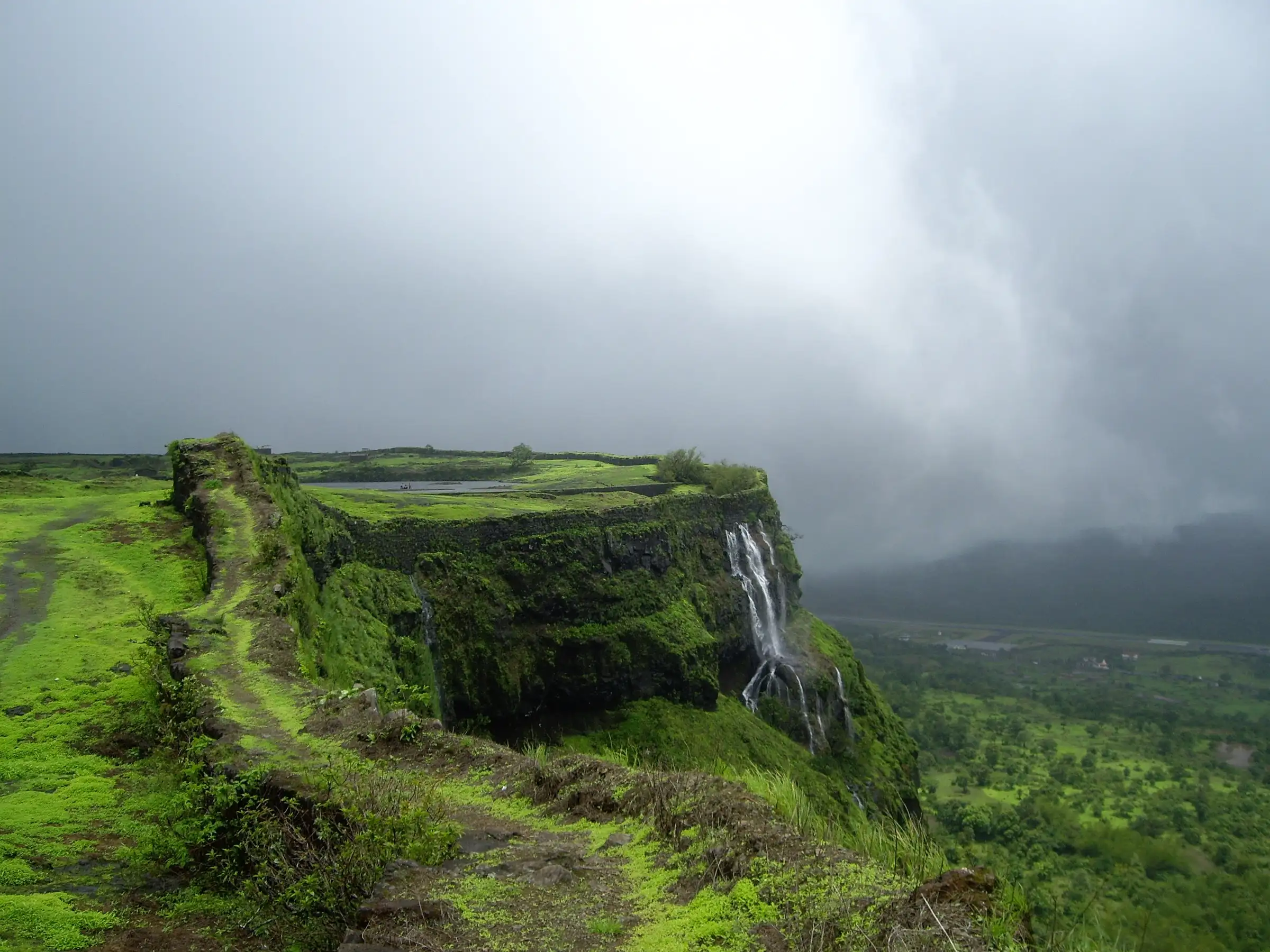 Green moss-covered rampart and bastion on the Korigad plateau, with a waterfall pouring off the cliff into the valley under monsoon cloud