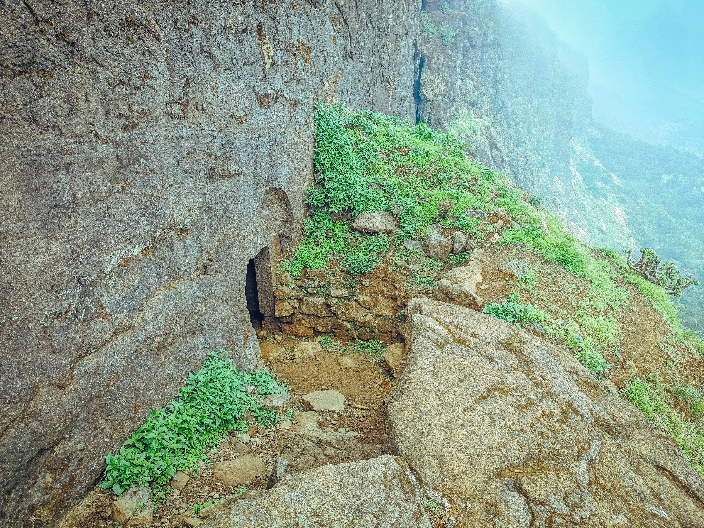 A rock-cut doorway at the foot of a basalt cliff on the Bhandardurg trail, with a stony path and green plants