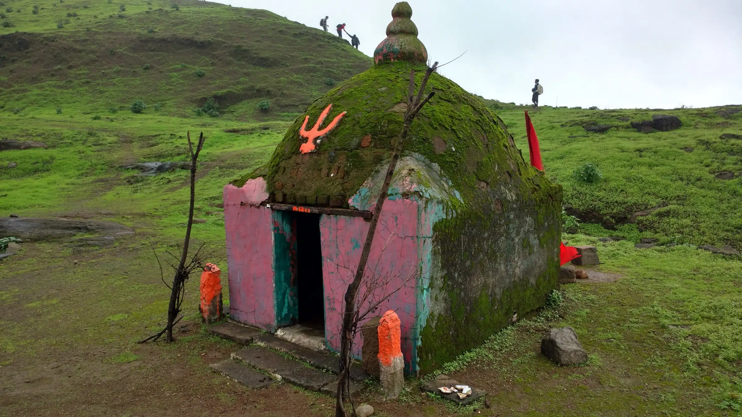 Small domed Bhavani Mata temple on the green top of Tringalwadi Fort, with trekkers on the slope behind