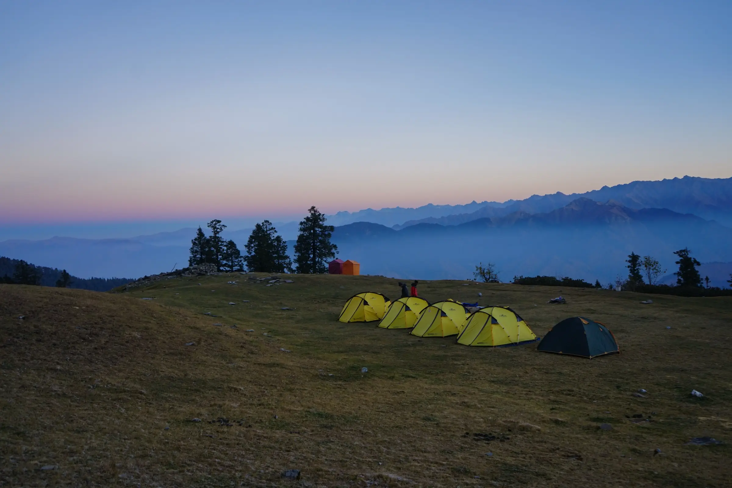 Yellow trekking tents on the grassy Bhoj Gadi campsite at dawn, with hazy mountain ranges behind