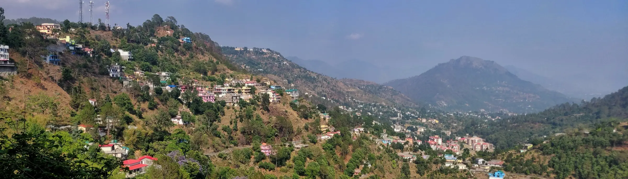Panorama of Bhowali town spread over forested hillsides, with hazy Kumaon hills beyond