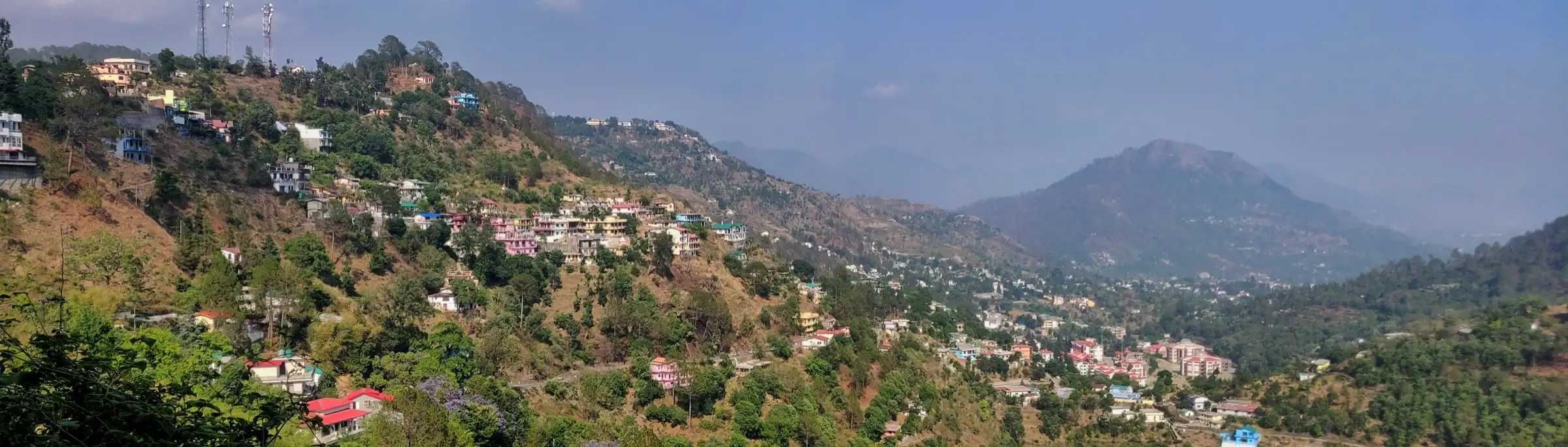 Panorama of Bhowali town spread over forested hillsides, with hazy Kumaon hills beyond