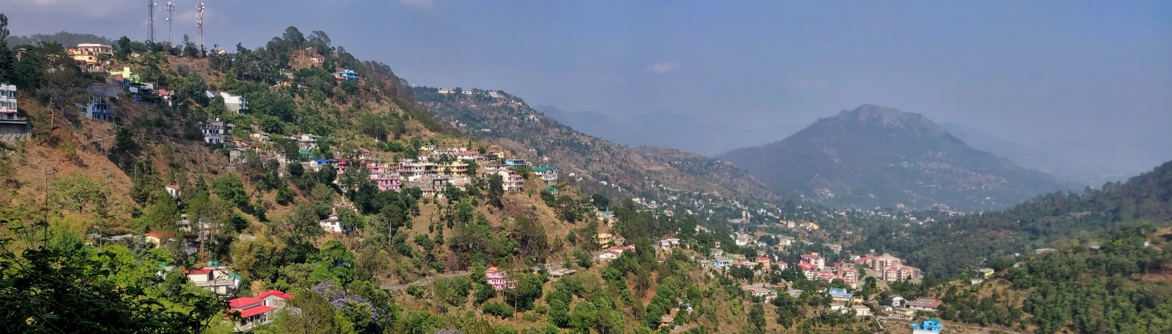 Panorama of Bhowali town spread over forested hillsides, with hazy Kumaon hills beyond