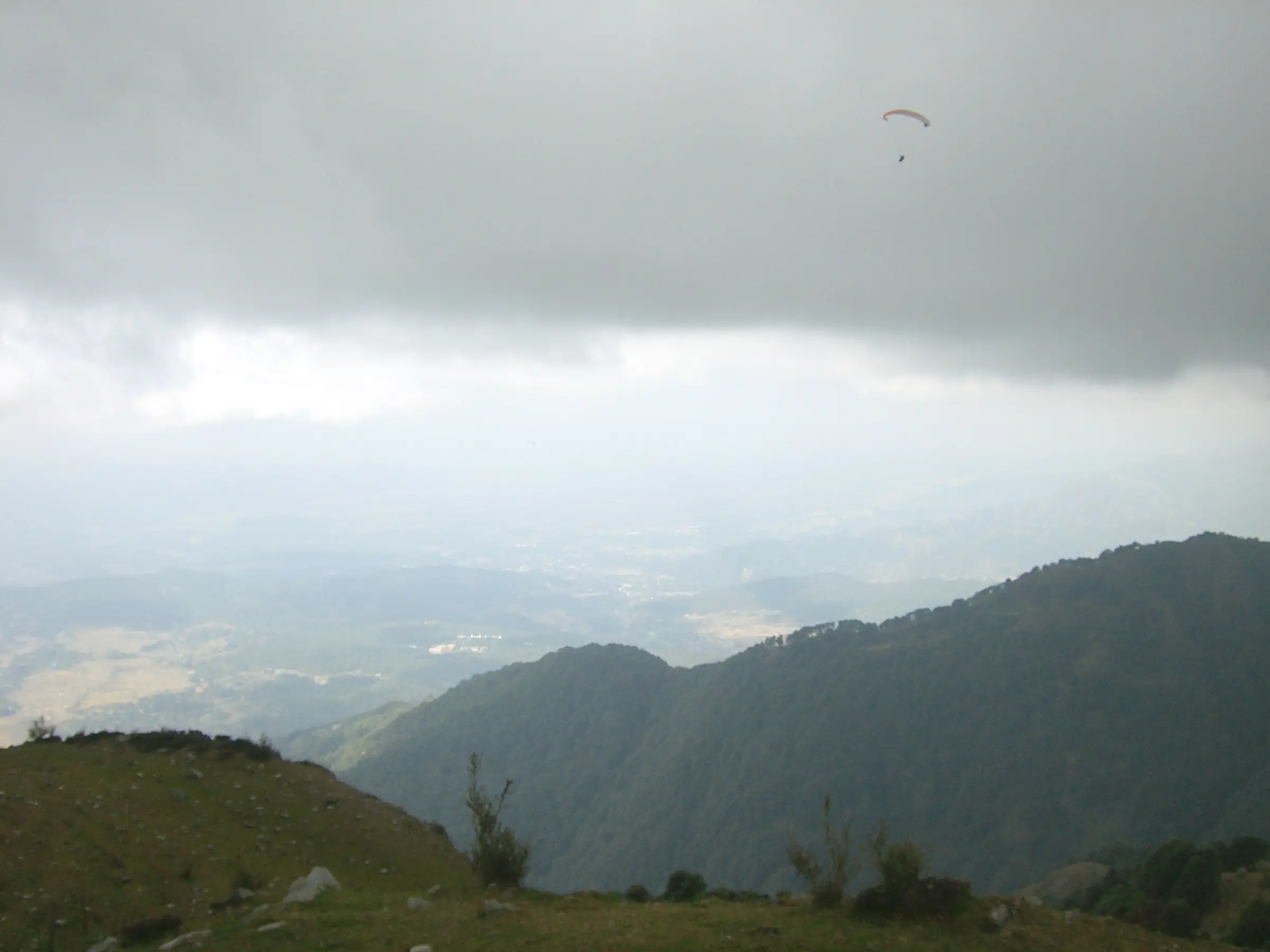 A paraglider over forested ridges and the Kangra valley seen from the grassy take-off at Billing, where the trek starts