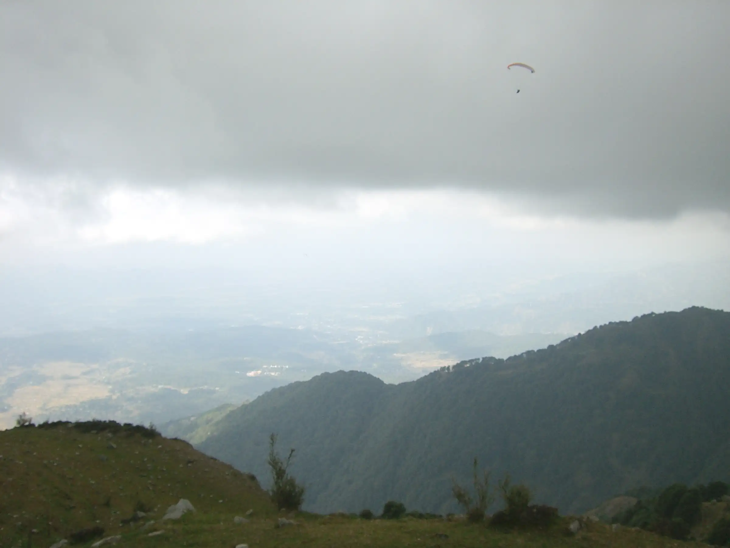 A paraglider over forested ridges and the Kangra valley seen from the grassy take-off at Billing, where the trek starts