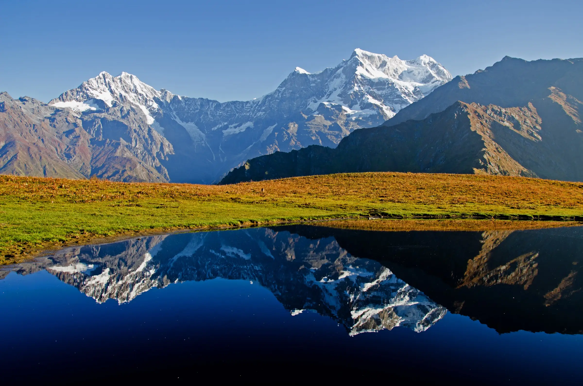 Snow-covered Chaukhamba massif reflected in a still pond on the autumn meadow at Buda Madmaheshwar under a clear blue sky