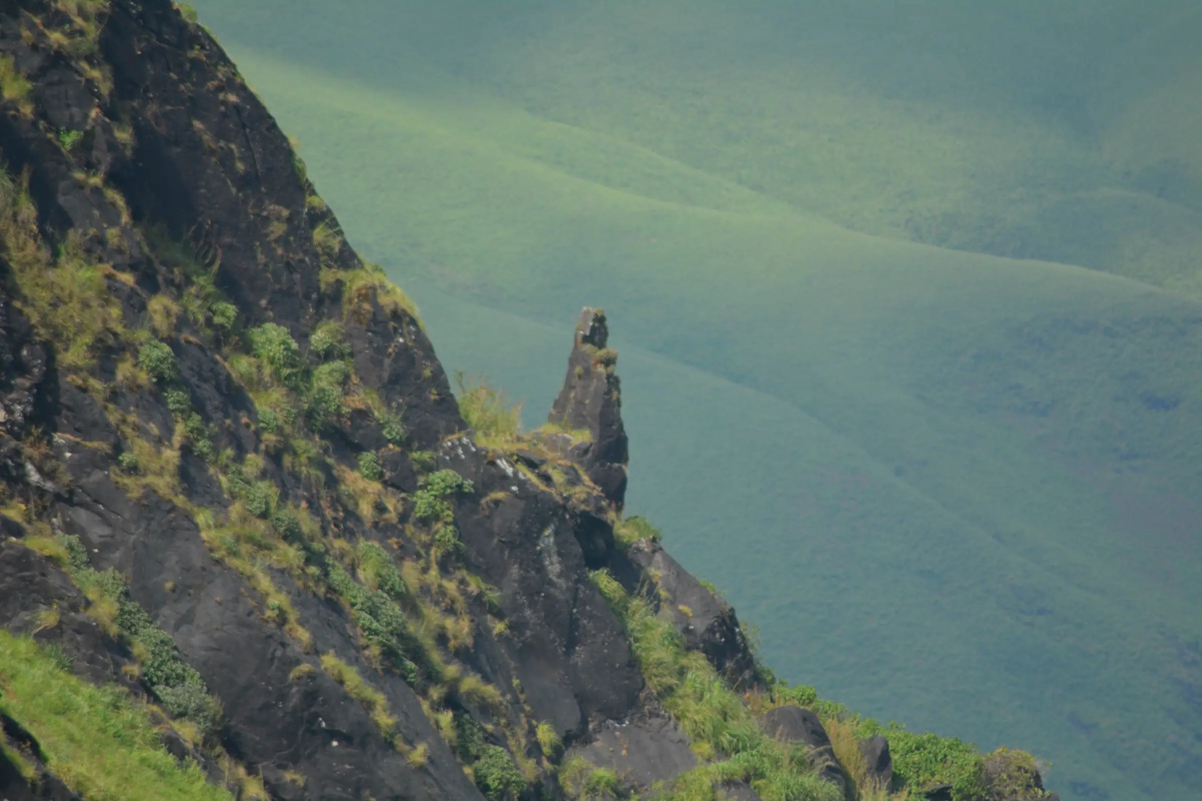 A lone rock pinnacle on a rocky grass-covered slope, seen on the way up Kudremukh, with green valley slopes behind