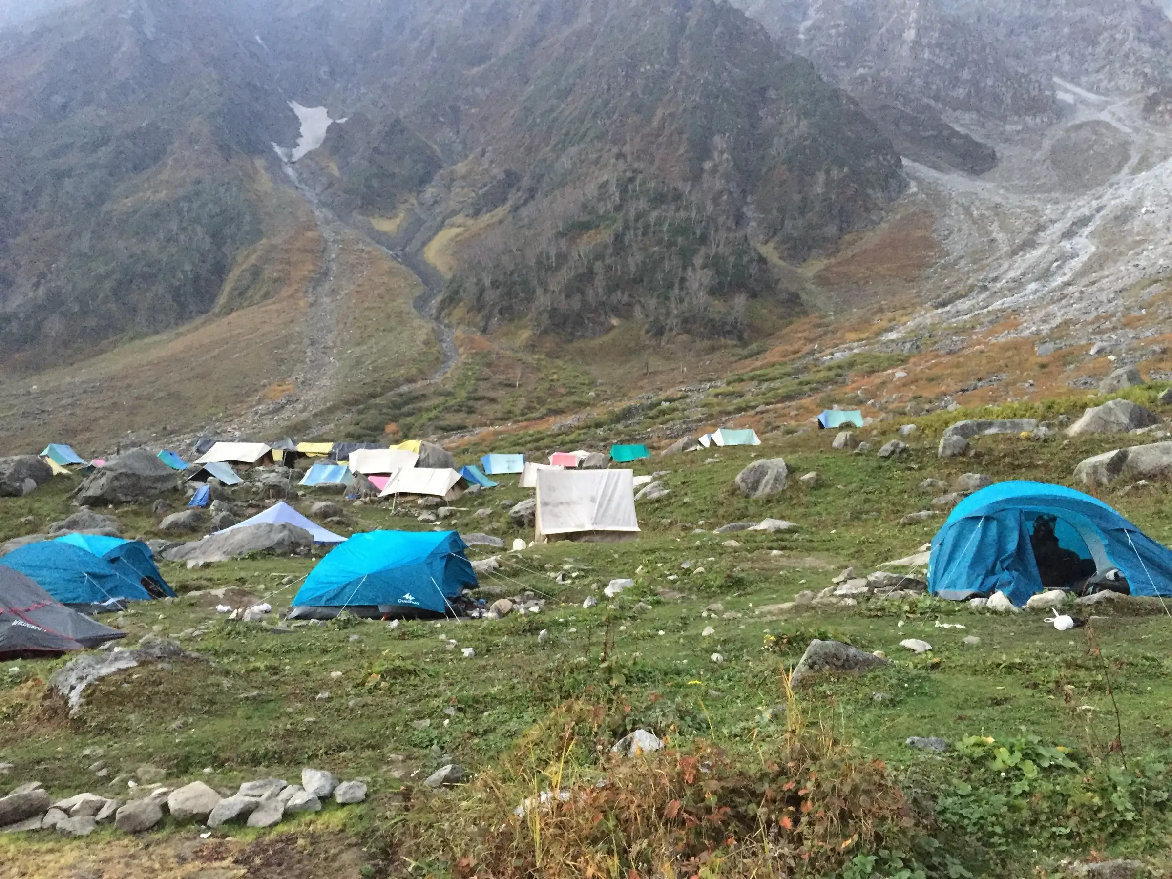 Blue and white trekking tents pitched on a grassy meadow below steep mountain slopes on the way to Beas Kund