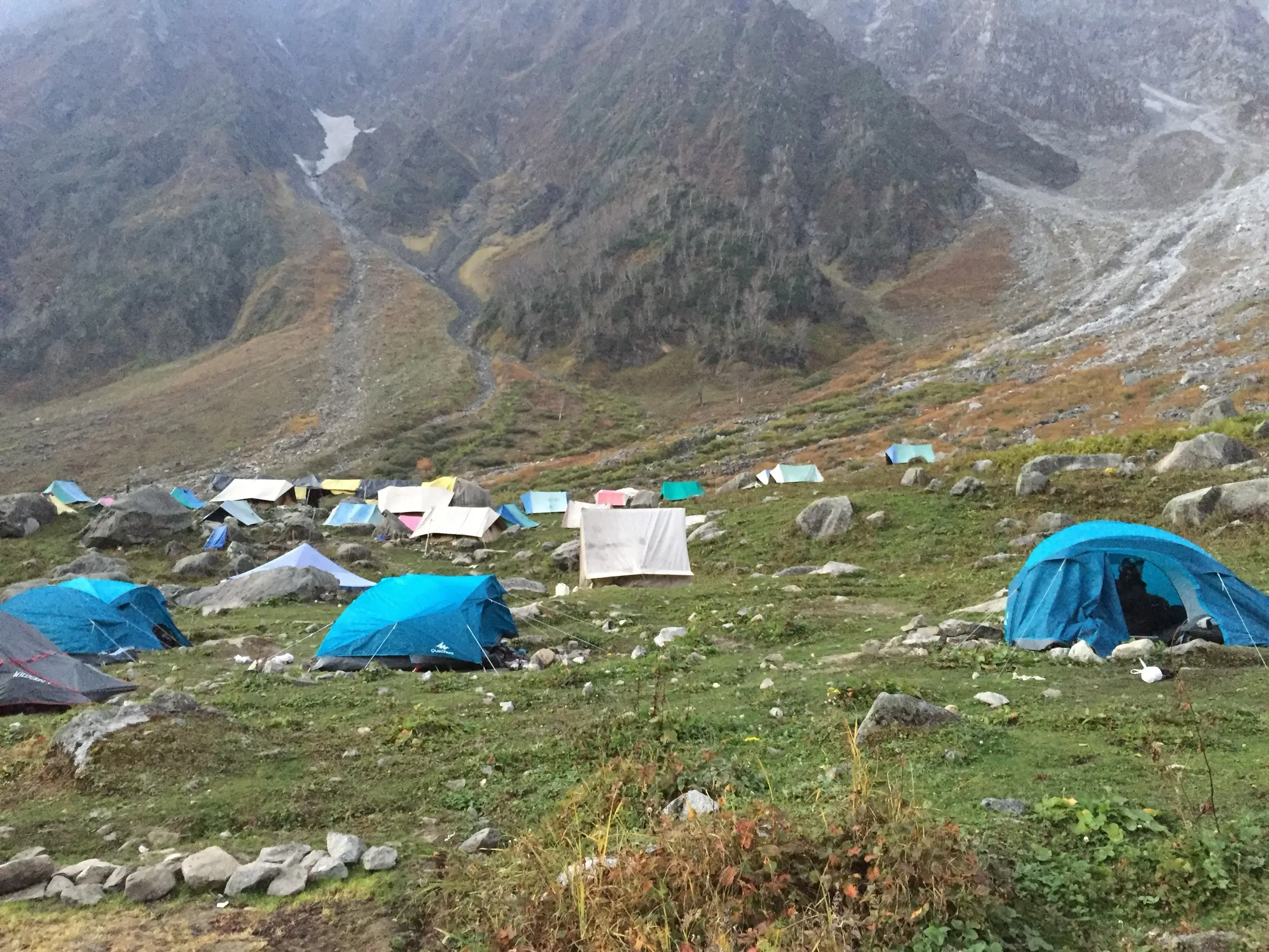 Blue and white trekking tents pitched on a grassy meadow below steep mountain slopes on the way to Beas Kund