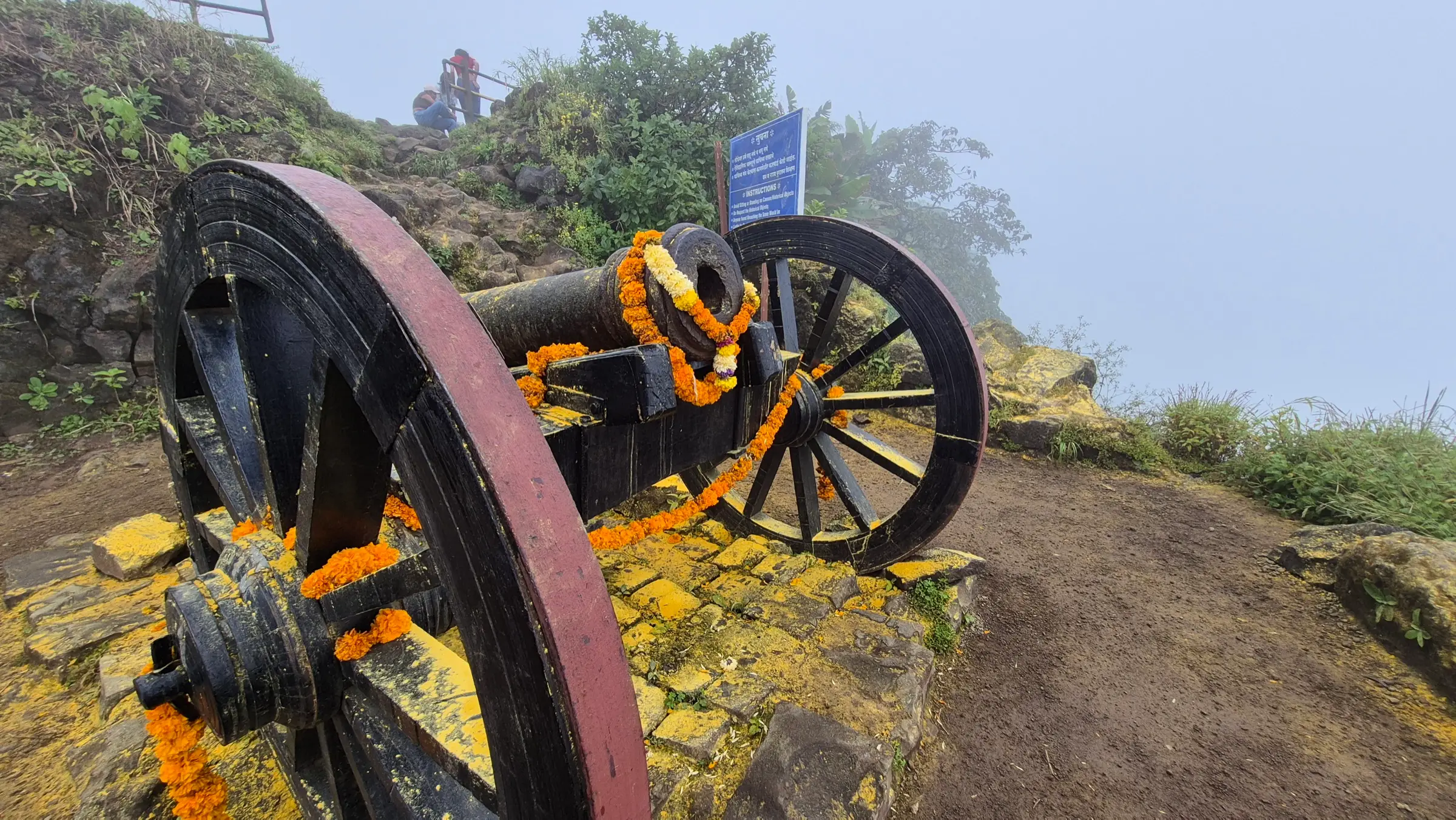 An old iron cannon on a wooden wheeled carriage, garlanded with marigolds, on a misty edge of Sinhagad Fort