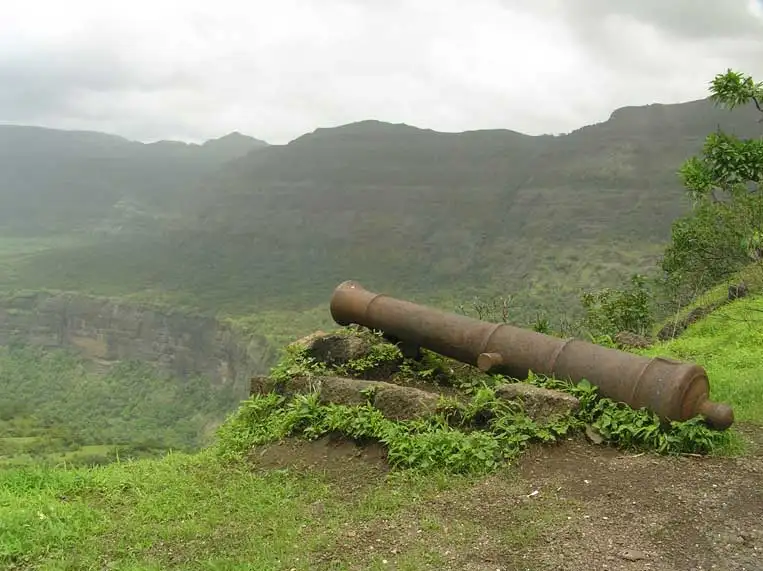 An old iron cannon resting on stones at Kothaligad, with green Sahyadri cliffs behind