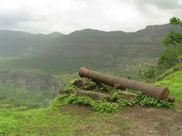 An old iron cannon resting on stones at Kothaligad, with green Sahyadri cliffs behind