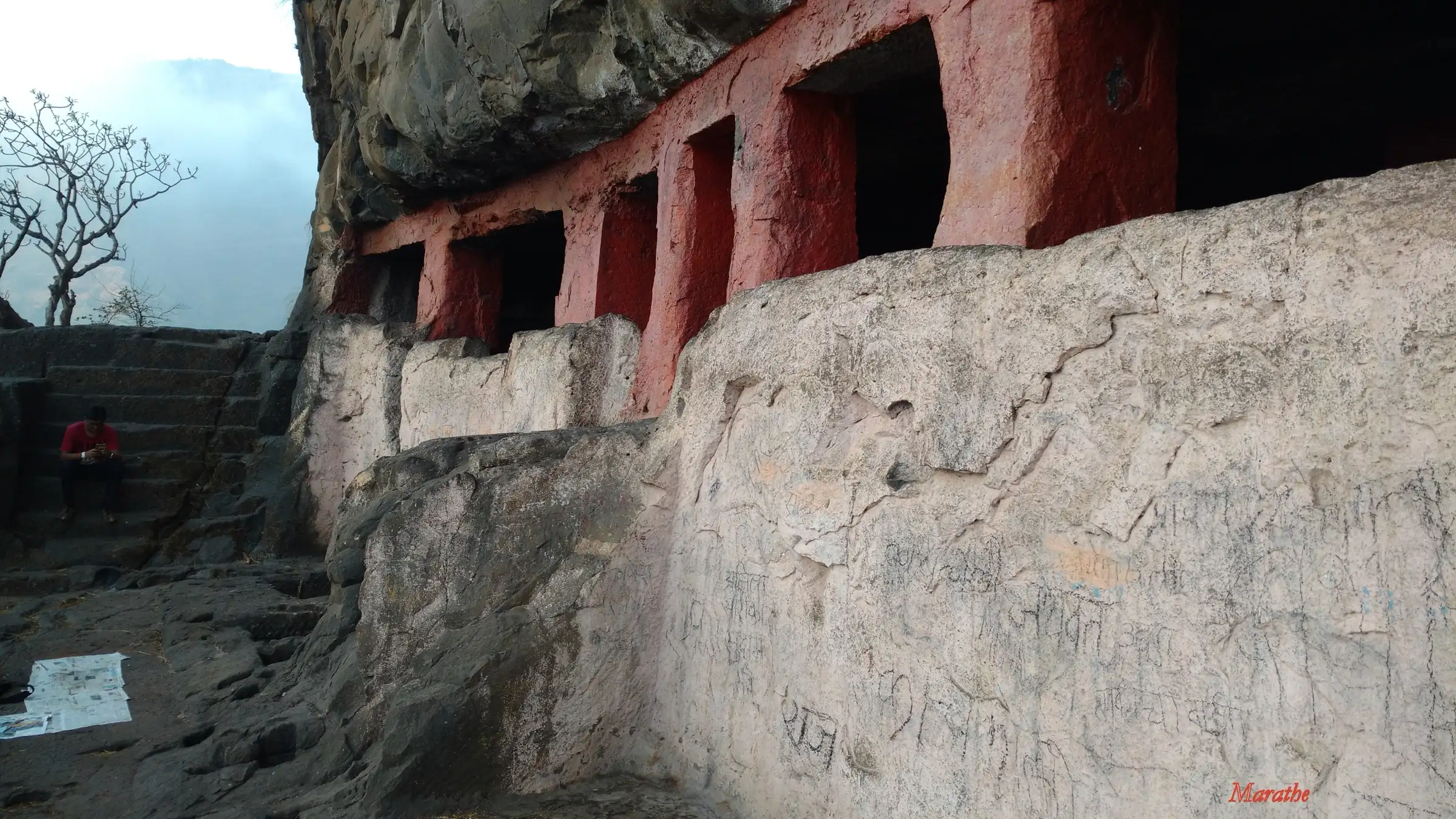 Rock-cut cave on Gorakhgad with square red-painted pillars under an overhanging cliff and steps to the left
