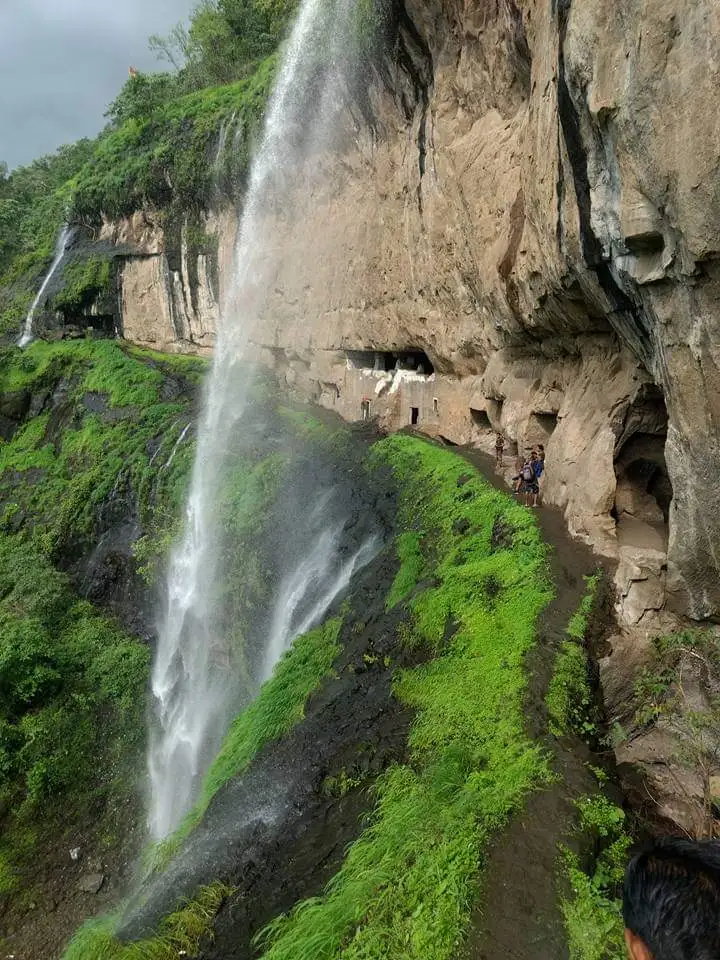 Ganpati Gadad caves cut into a tall rock cliff, with a waterfall plunging past a grassy ledge where trekkers stand by the cave mouths