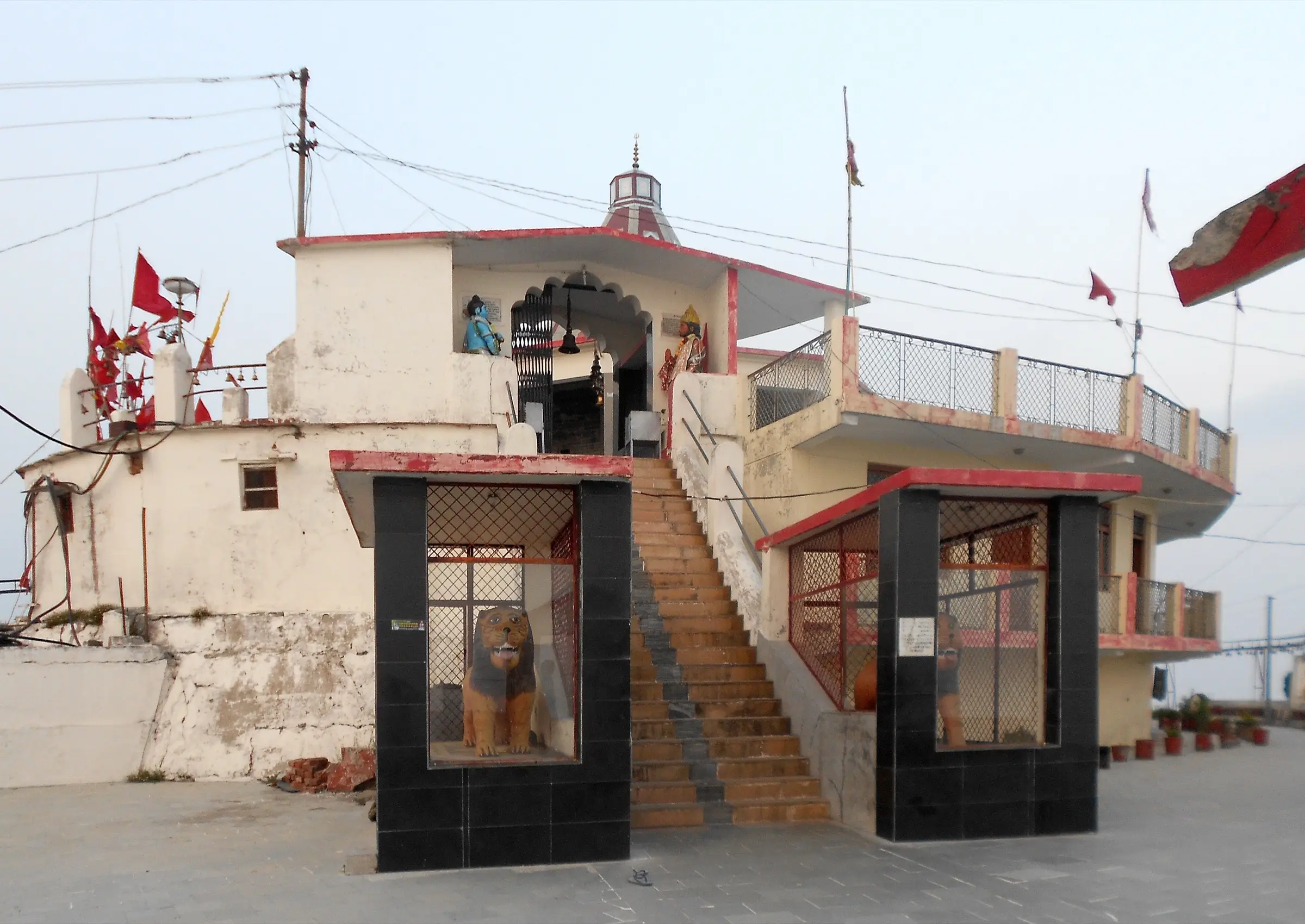 Entrance of Chandrabadni temple: stone steps rising between two lion statues in black-tiled shrines, red flags on the white temple walls
