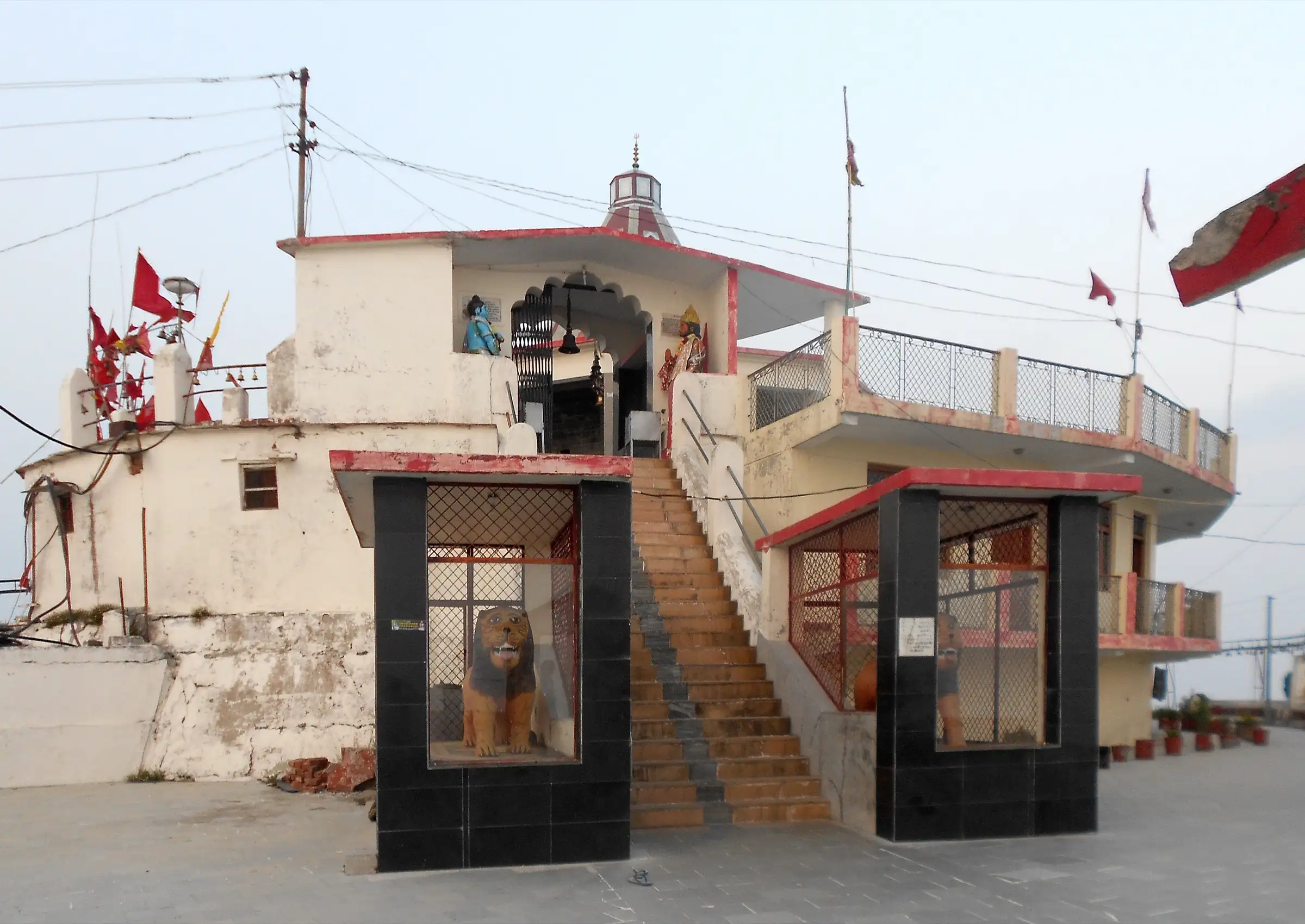 Entrance of Chandrabadni temple: stone steps rising between two lion statues in black-tiled shrines, red flags on the white temple walls
