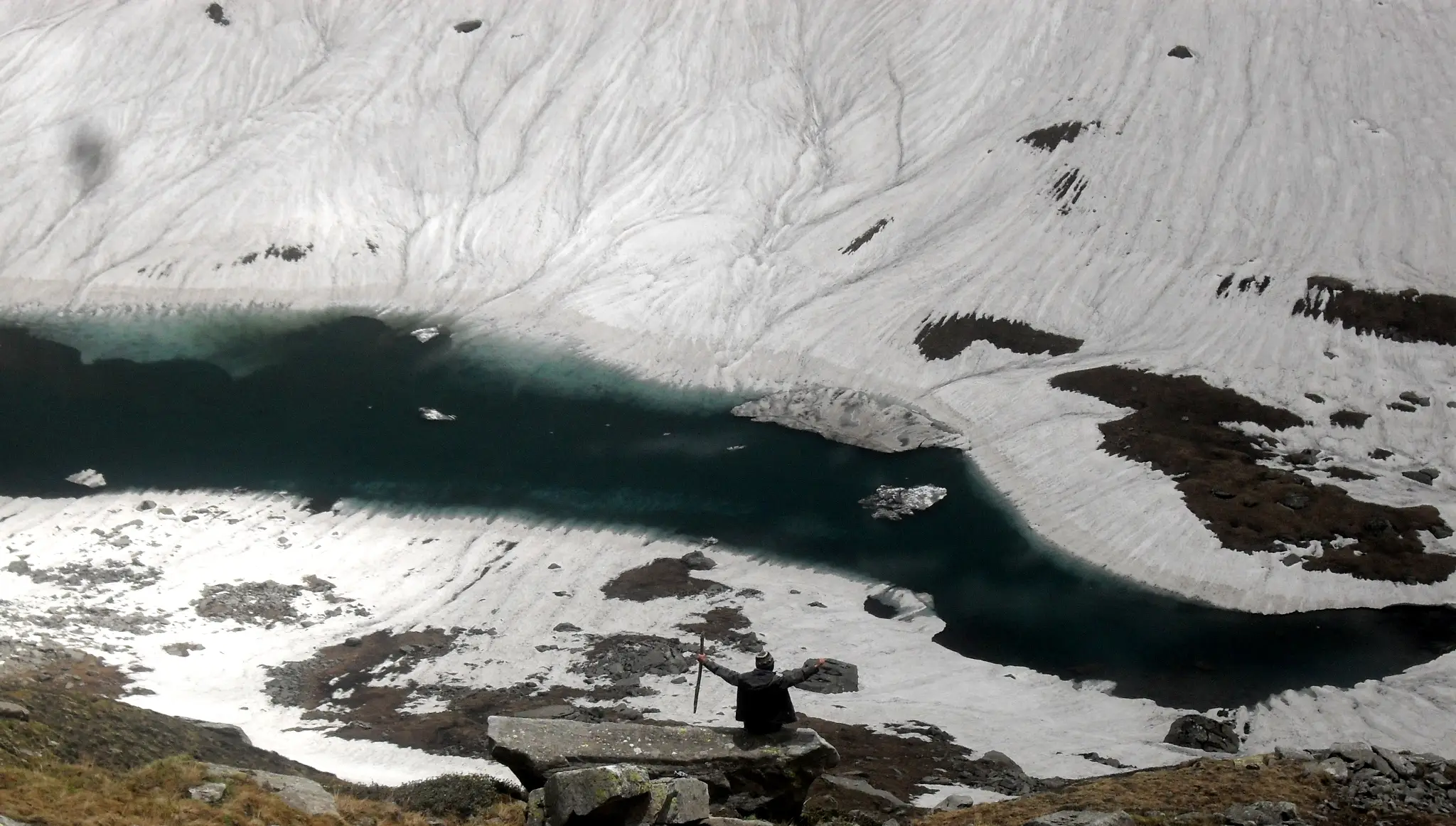 A trekker with arms raised on a rock above a dark, partly frozen Chandranahan lake ringed by snowfields