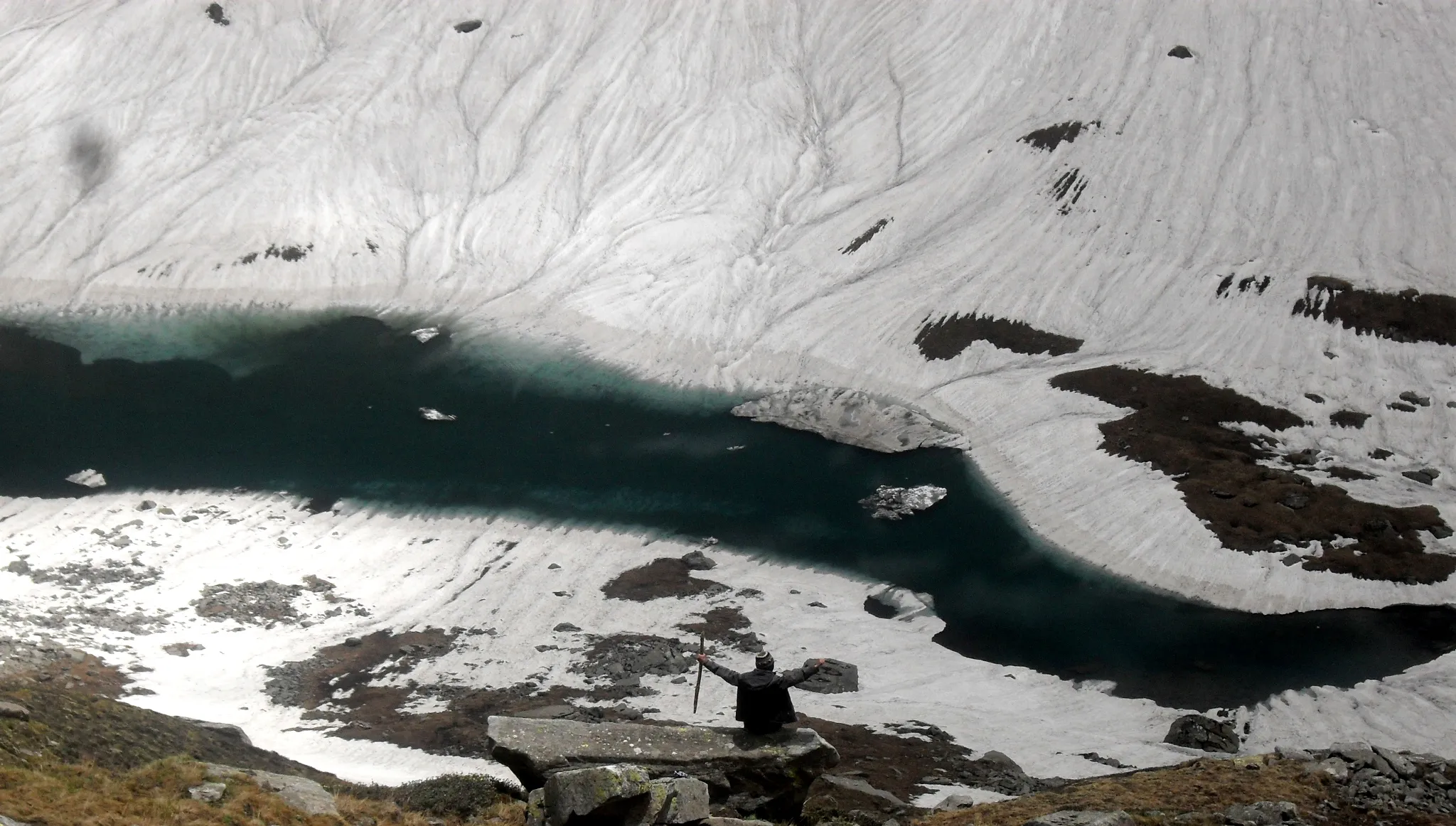 A trekker with arms raised on a rock above a dark, partly frozen Chandranahan lake ringed by snowfields