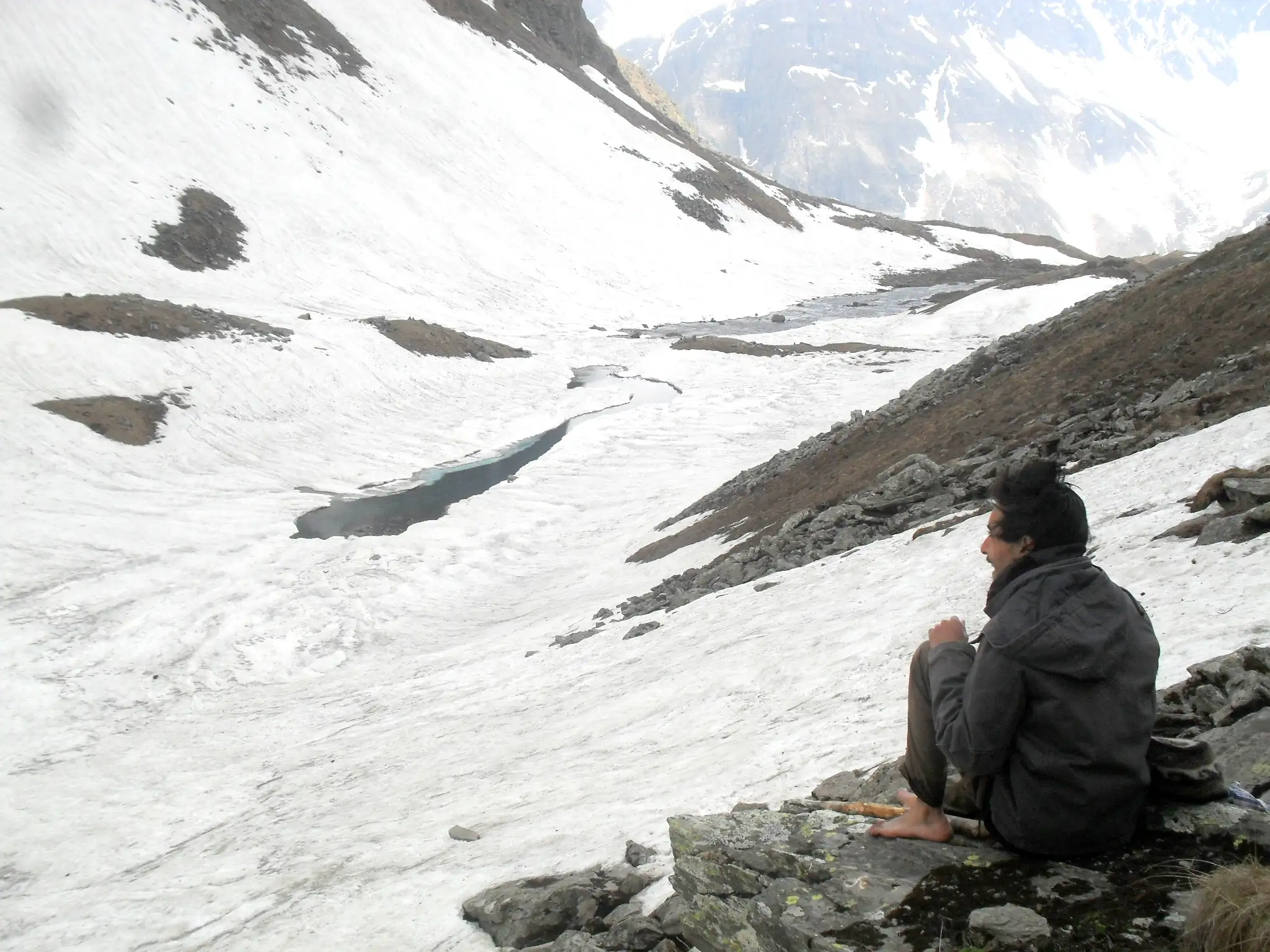 A trekker sitting on a rock above a partly frozen Chandranahan lake in a snow-filled bowl