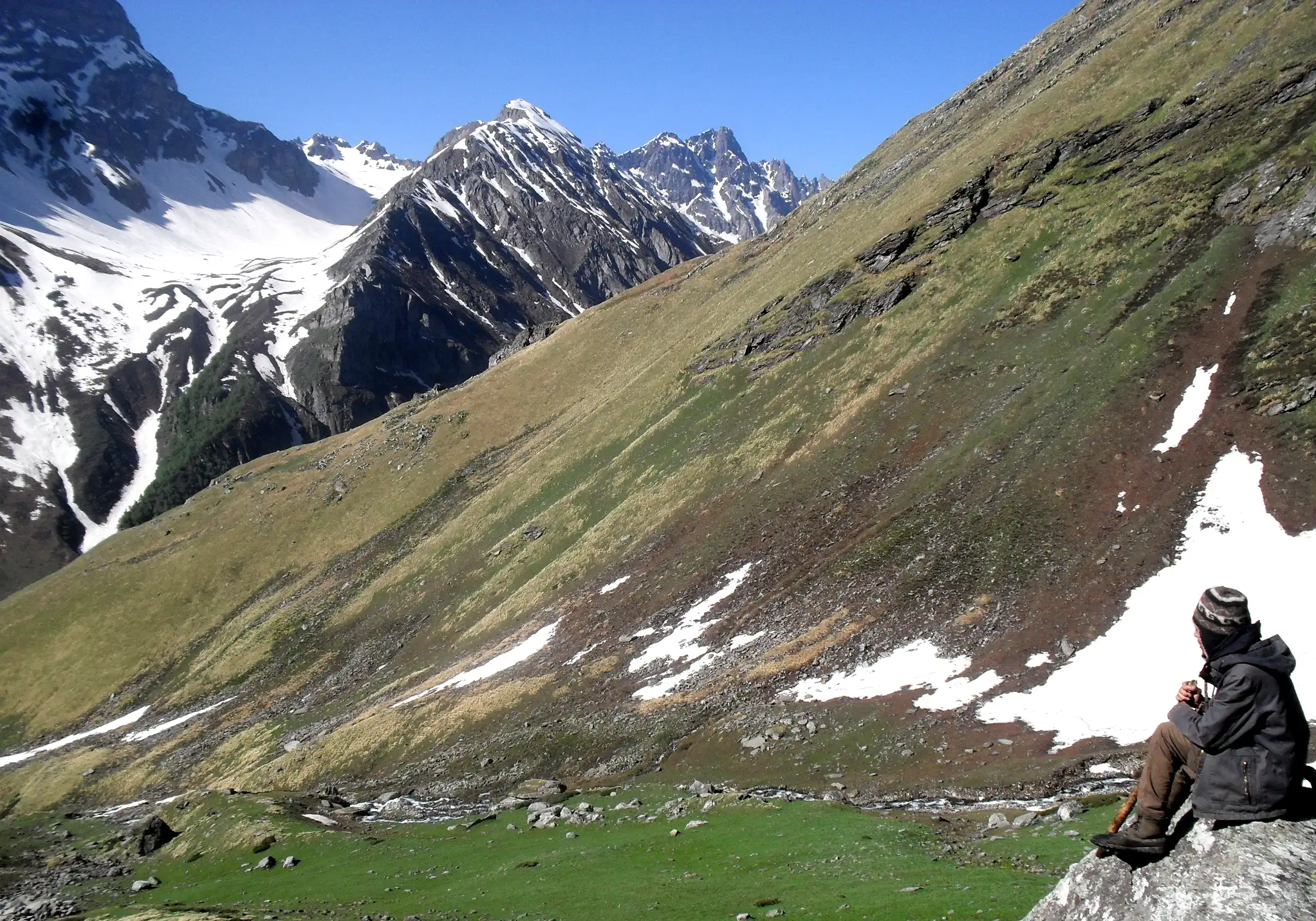 A trekker sitting on a rock above a green meadow with snow patches and snowy peaks in the Chandranahan area