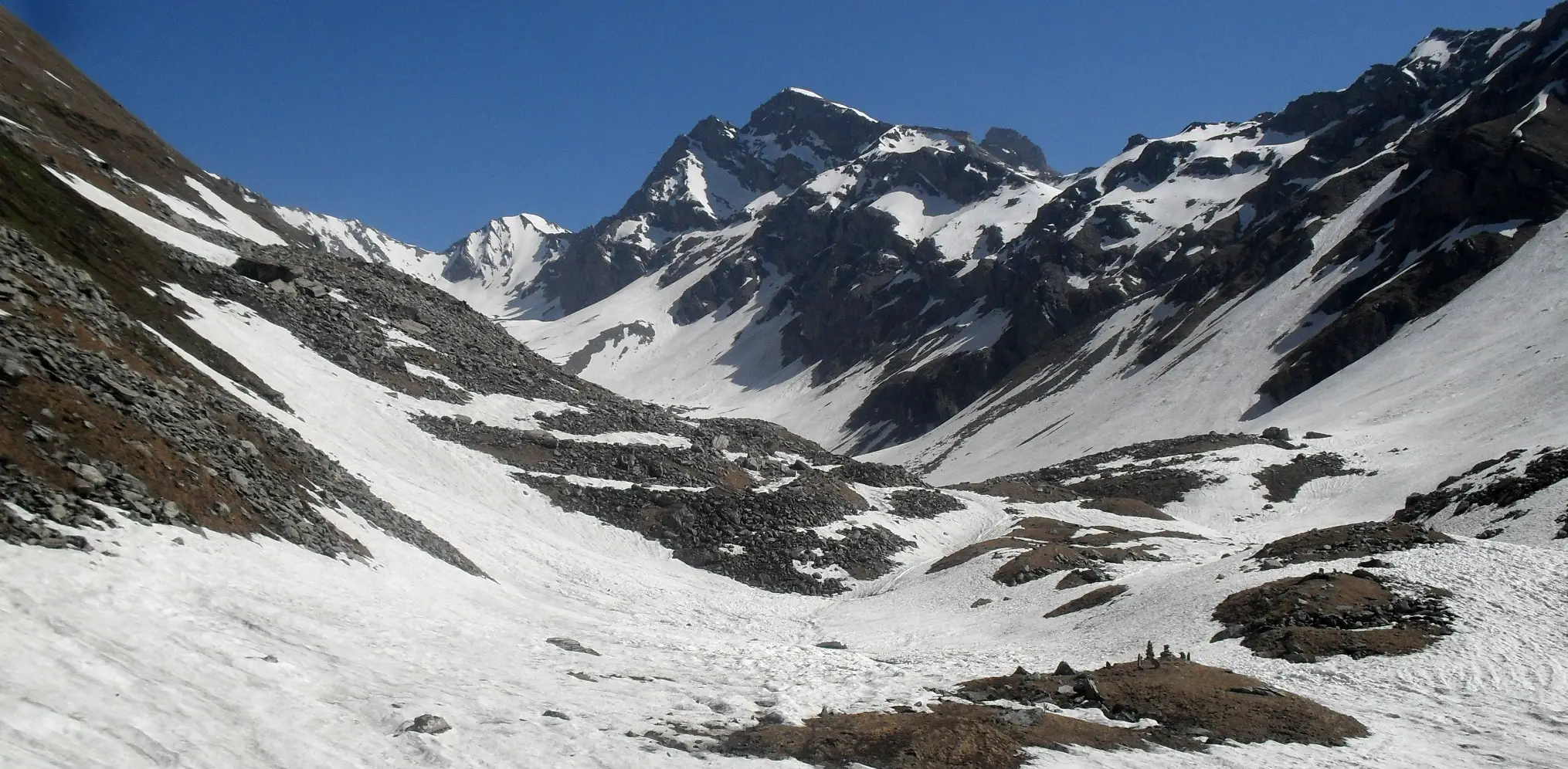 Snowfields and boulders in the Chandranahan bowl below jagged snow-streaked rock peaks under a blue sky