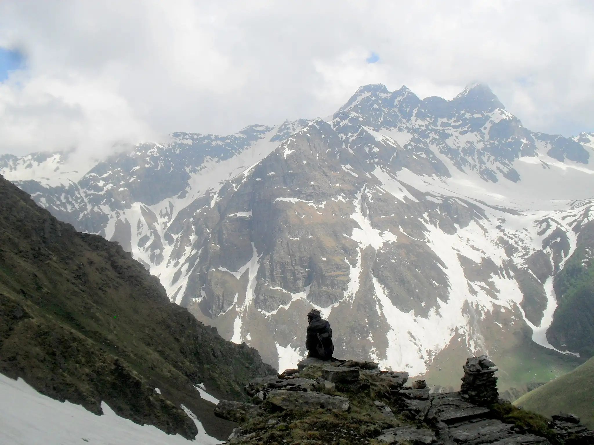 A trekker resting by a stone cairn facing snow-streaked rock peaks in the Chandranahan area