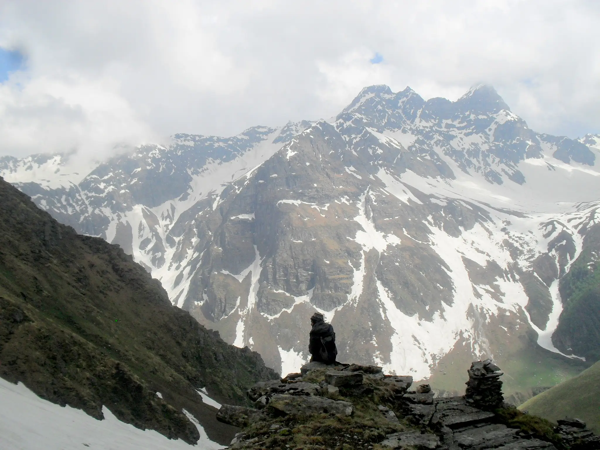 A trekker resting by a stone cairn facing snow-streaked rock peaks in the Chandranahan area