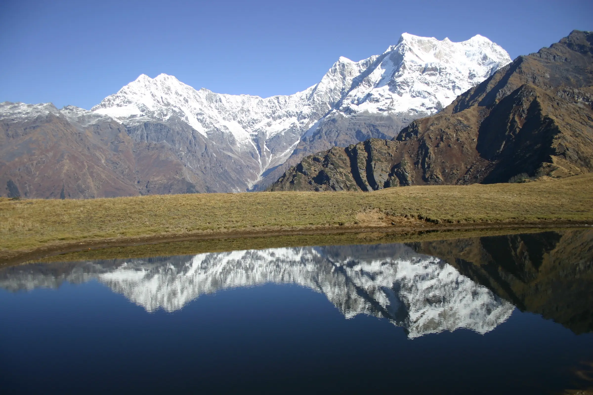 Snow-covered Chaukhamba massif reflected in a still pond on the grassy meadow above Madmaheshwar, the start of the climb towards Nandi Kund