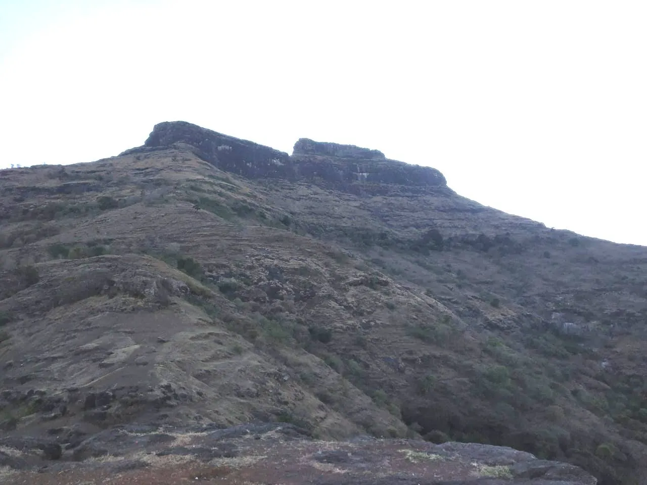 The flat-topped rock scarp of Chaulher Fort rising above dry, terraced slopes, seen from the hill above Wadi Chaulher