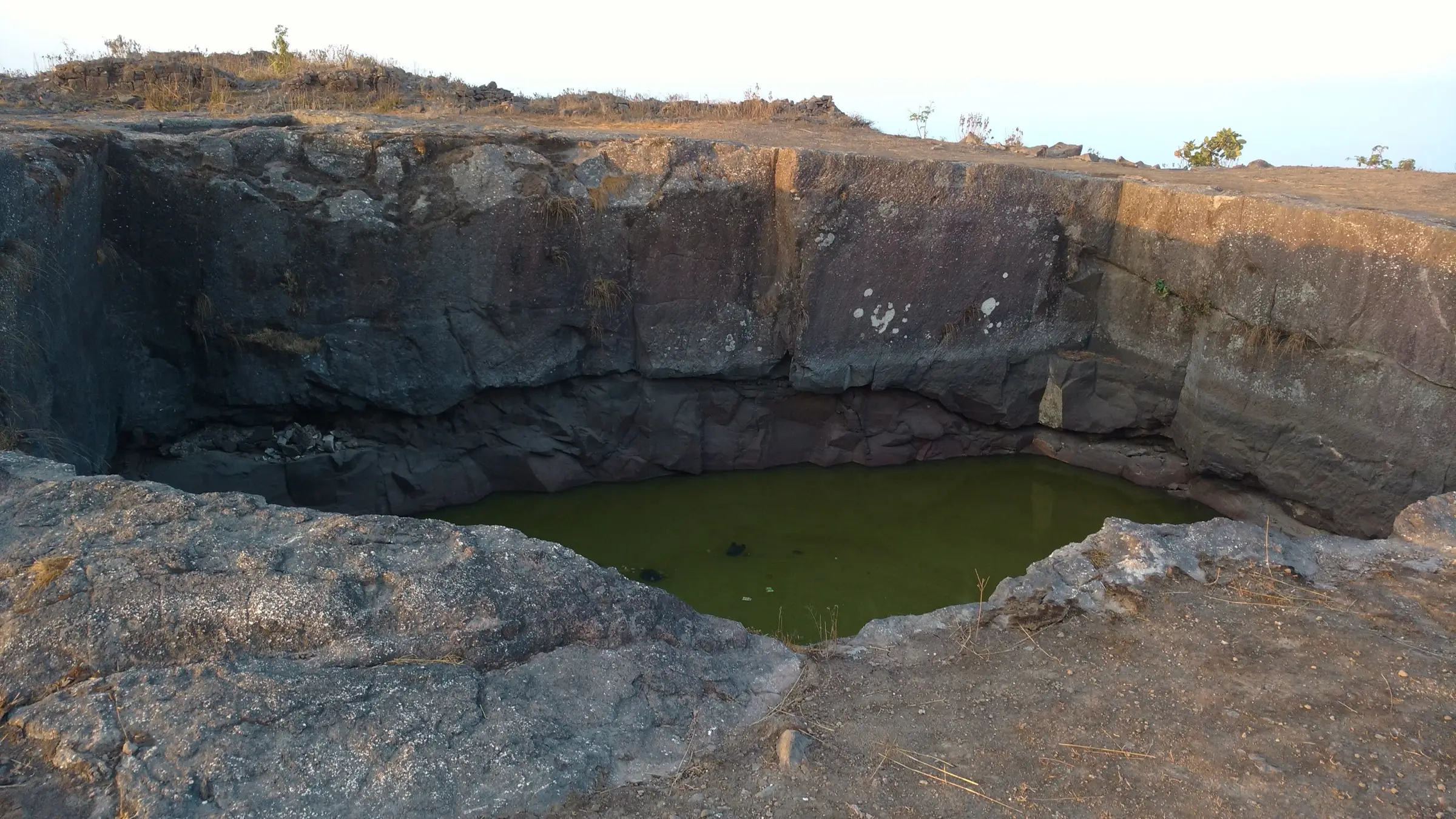 Large rock-cut water cistern with green water on top of Manikgad fort