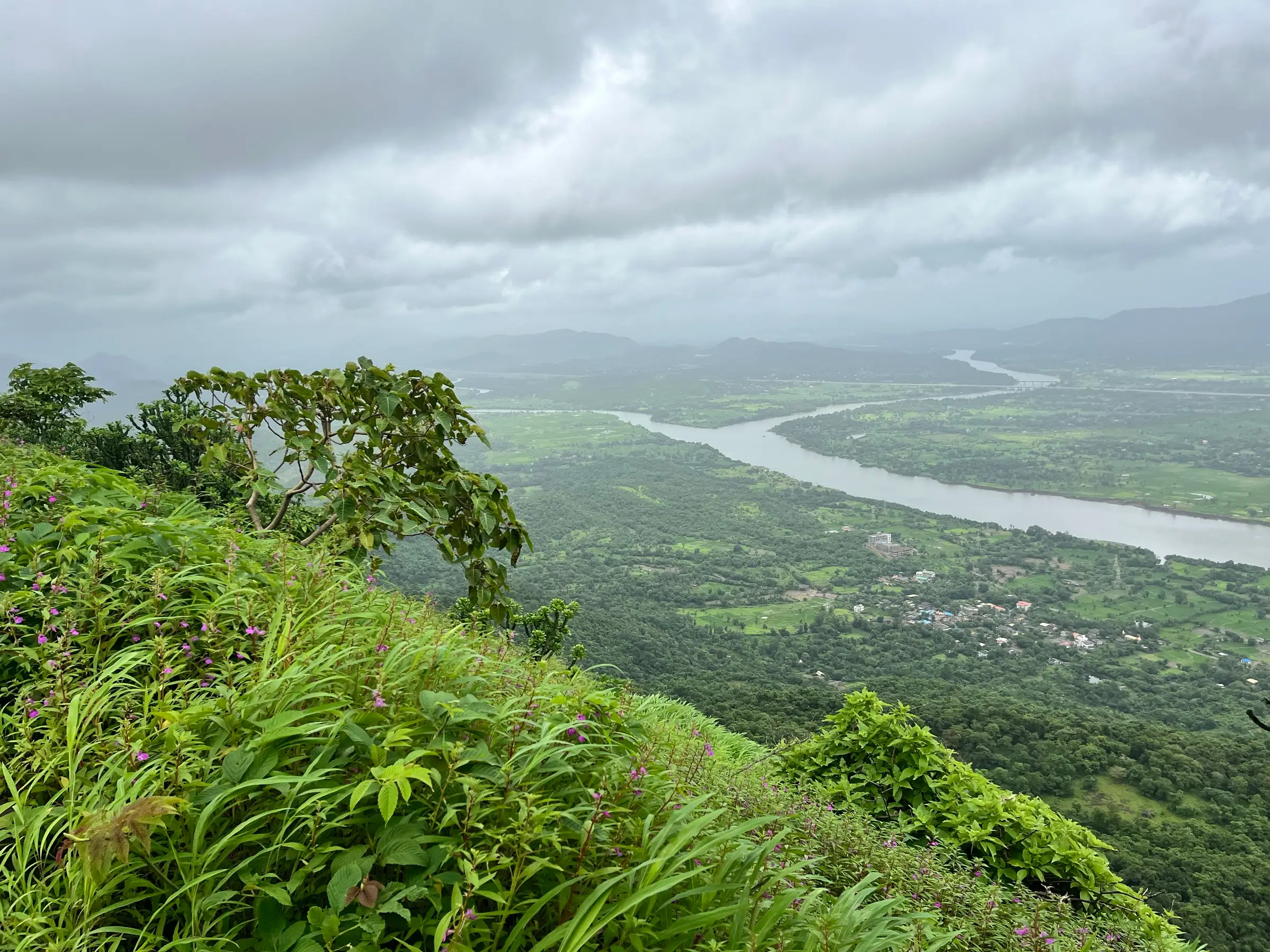 The Vaitarna river winding through green fields and villages below, seen from the grassy slopes of Tandulwadi Fort