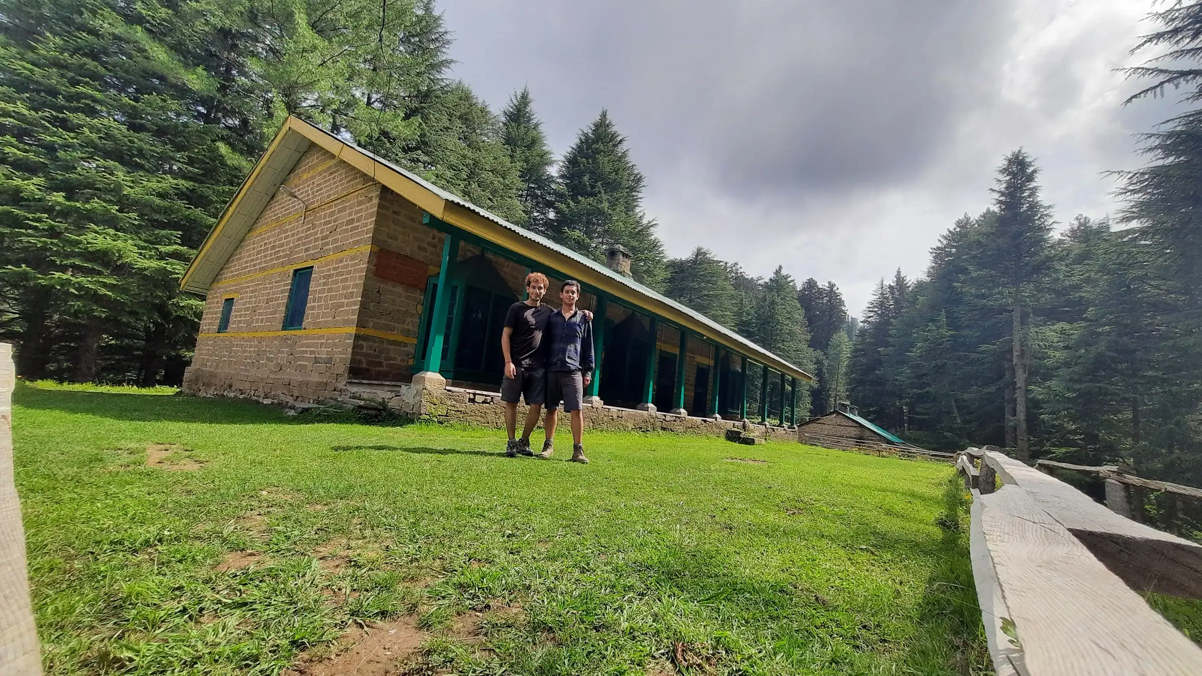 Two hikers in front of the long stone Kaisdhar Forest Rest House on a lawn surrounded by deodar forest
