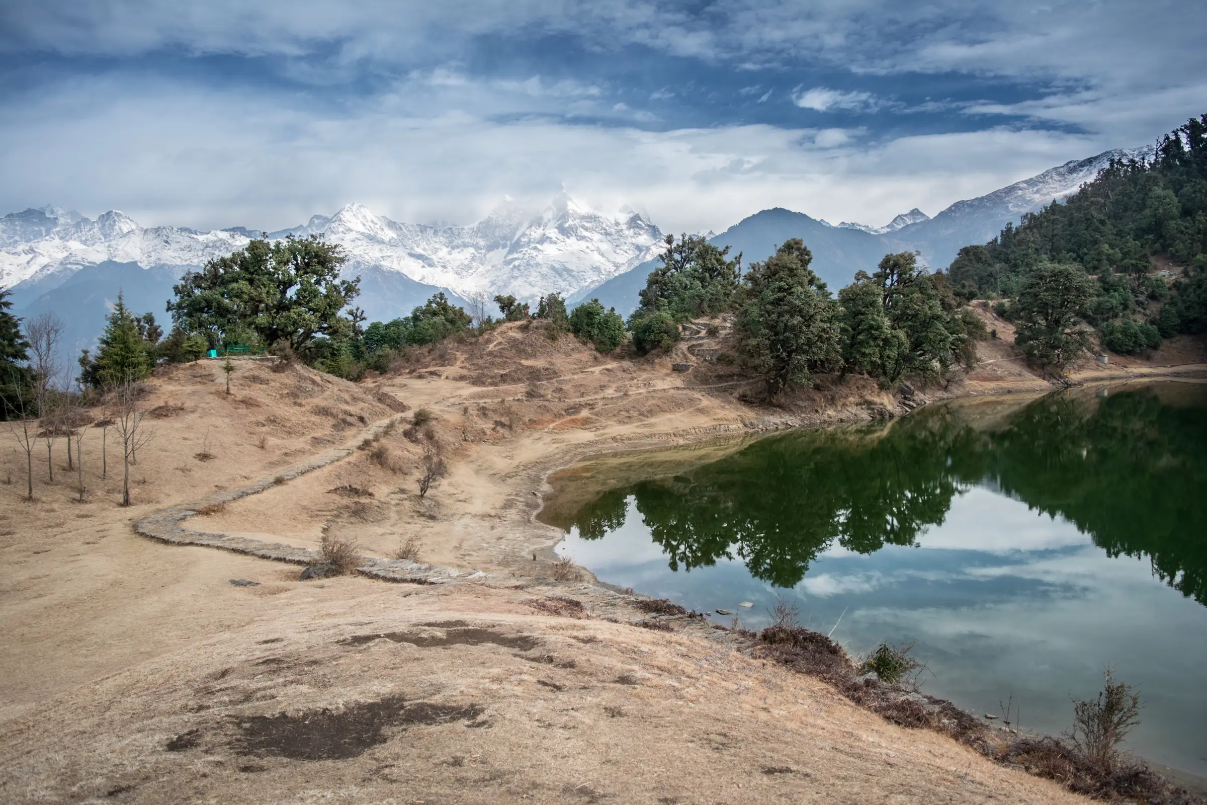 Deoria Tal lake reflecting oak trees, with a stone-edged path along its shore and snowy Chaukhamba peaks behind