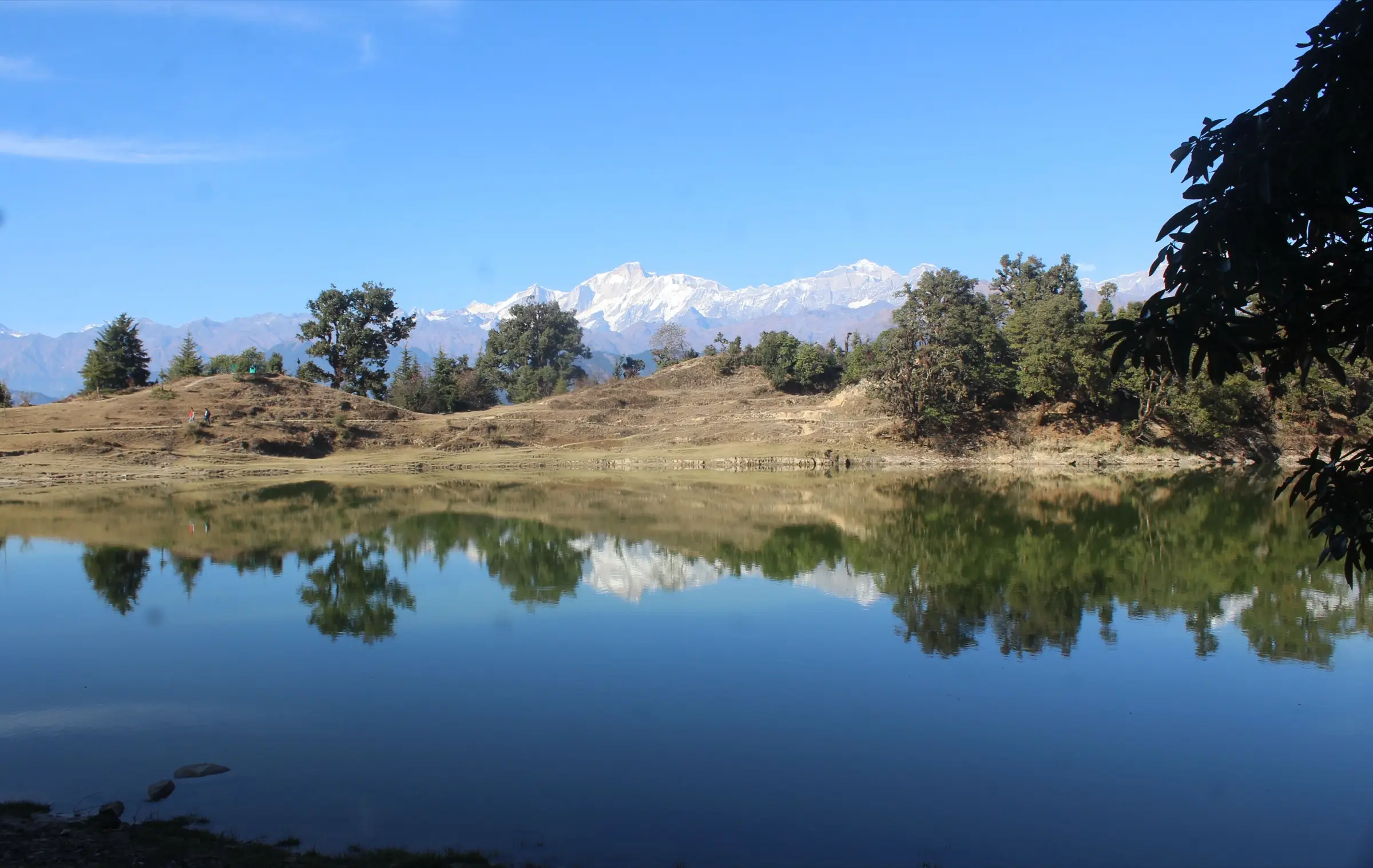 Still water of Deoriatal reflecting oak trees and a snowy Himalayan range under a clear blue sky