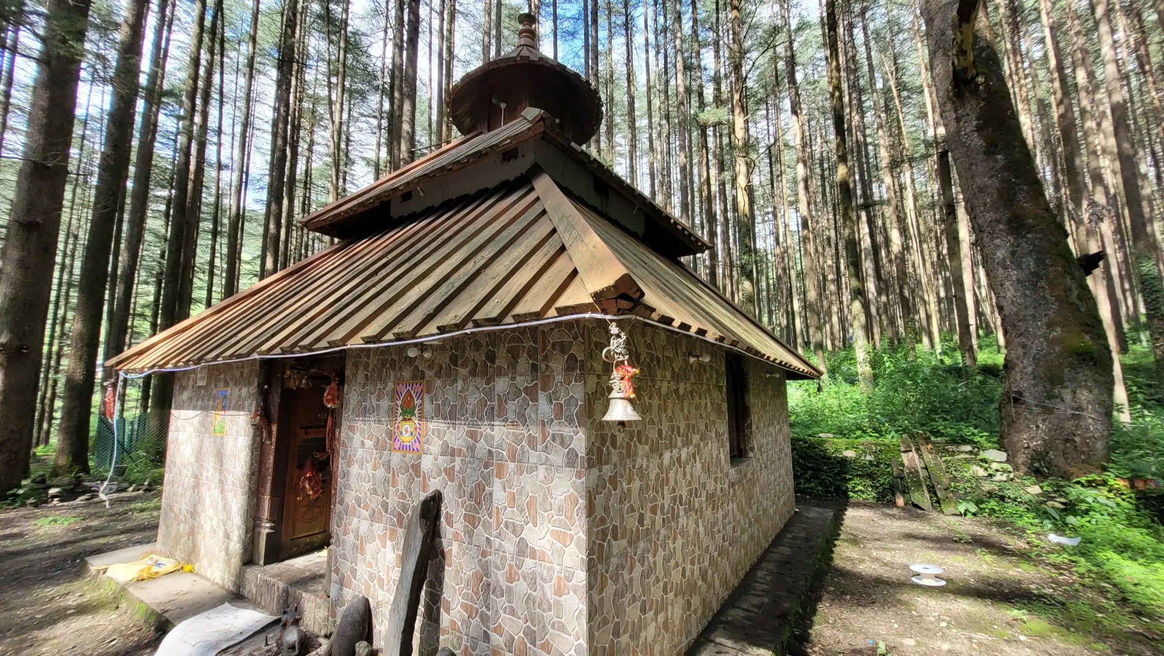 Small stone-walled Devalsari Shiva temple with a wooden roof and brass bell among tall deodar trees