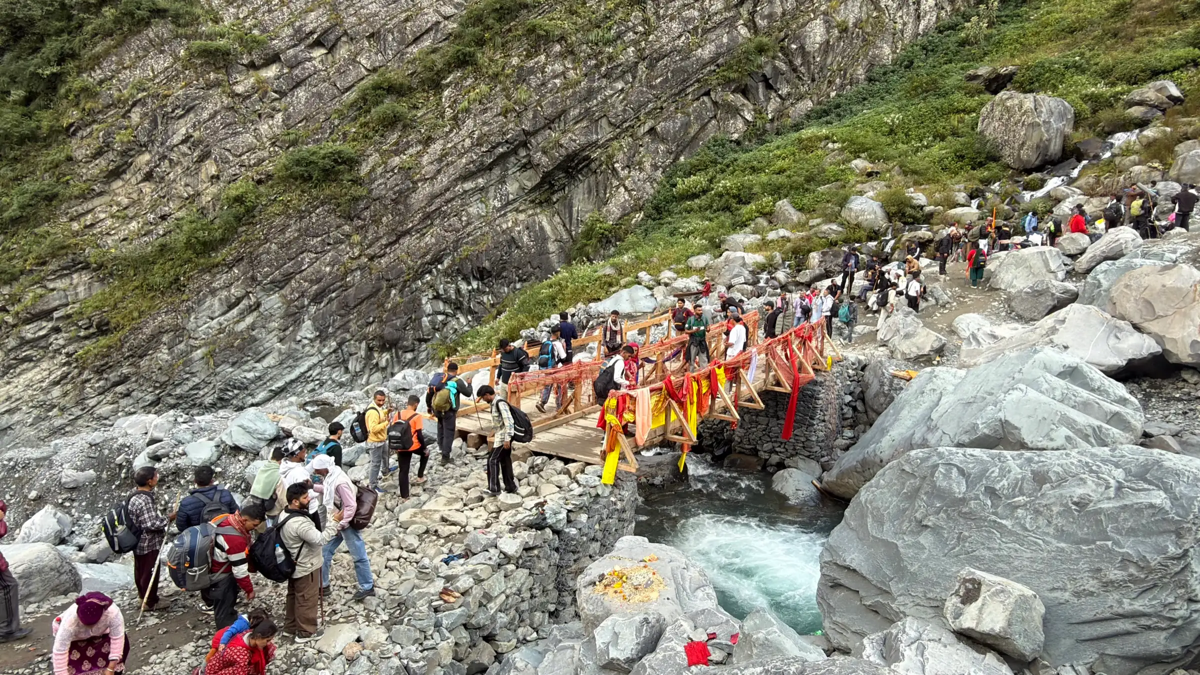 Pilgrims crossing a wooden bridge hung with red and yellow cloth over a rushing stream at Dhancho