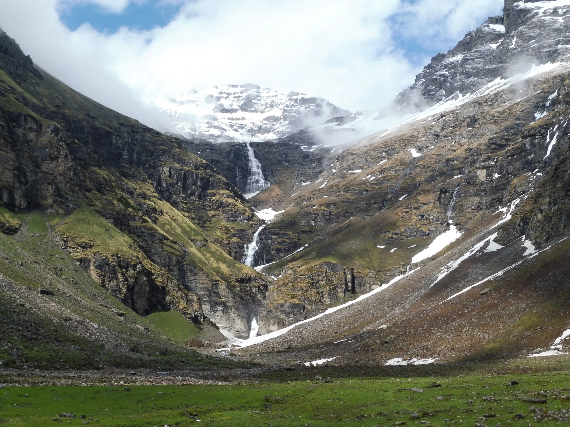 Green meadow of Dhanderas Thatch below the tall Rupin waterfall falling down a rocky cliff, with snowy peaks in cloud above